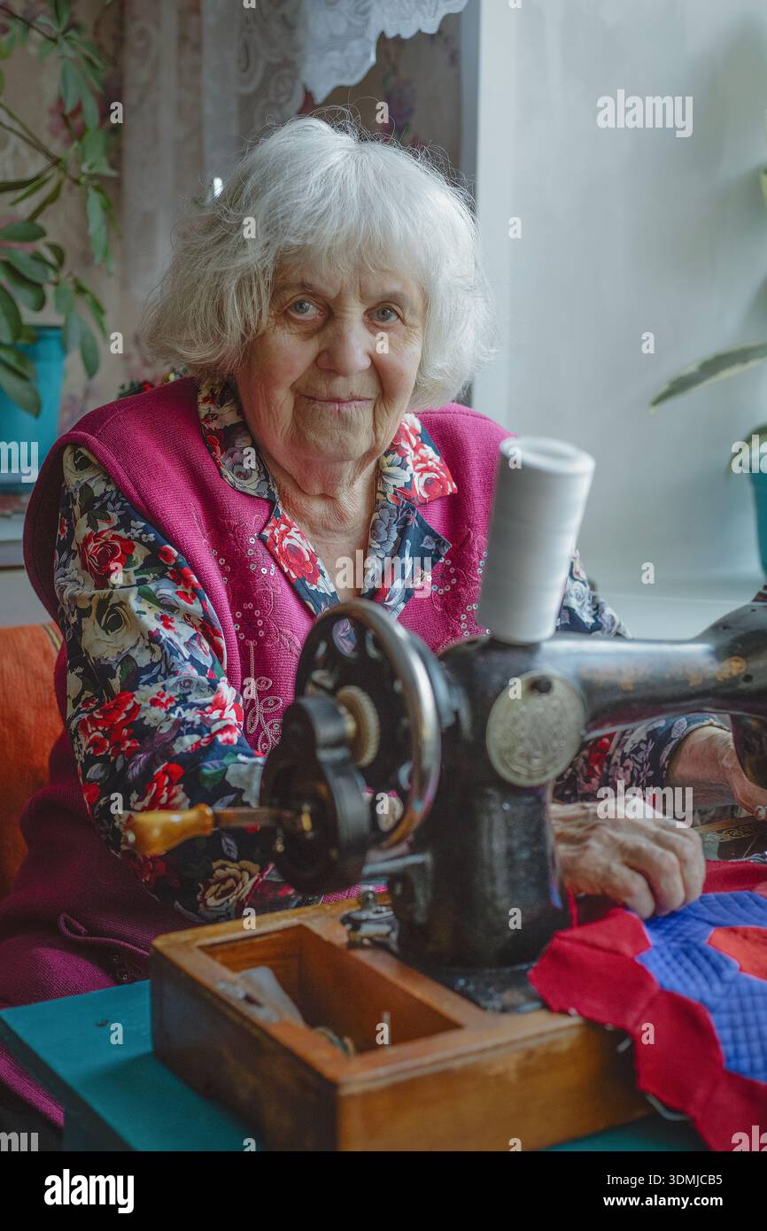 An elderly woman looks at the camera while using a sewing machine Stock Photo