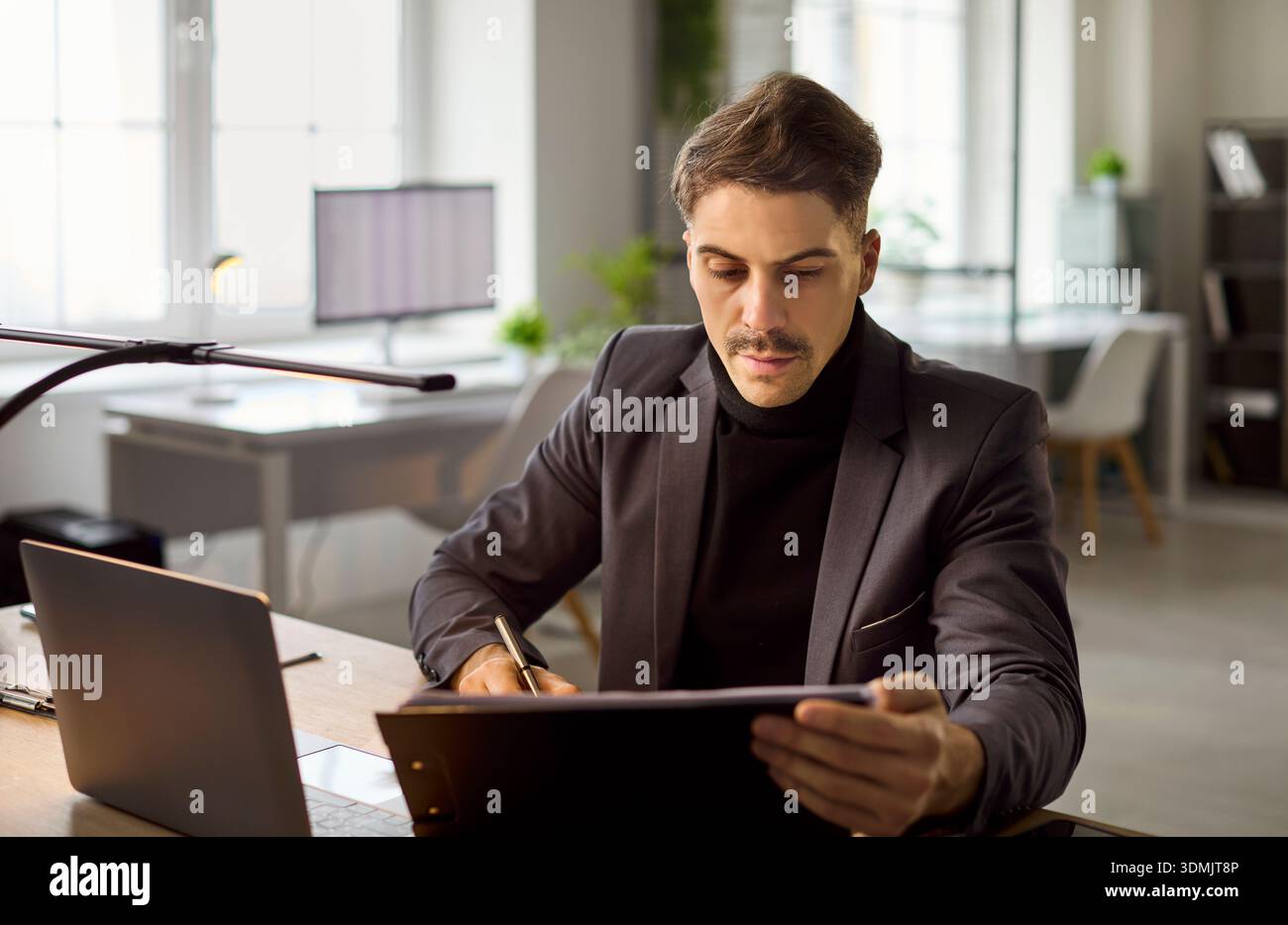 Focused businessman in suit reviewing documents at office desk with laptop Stock Photo