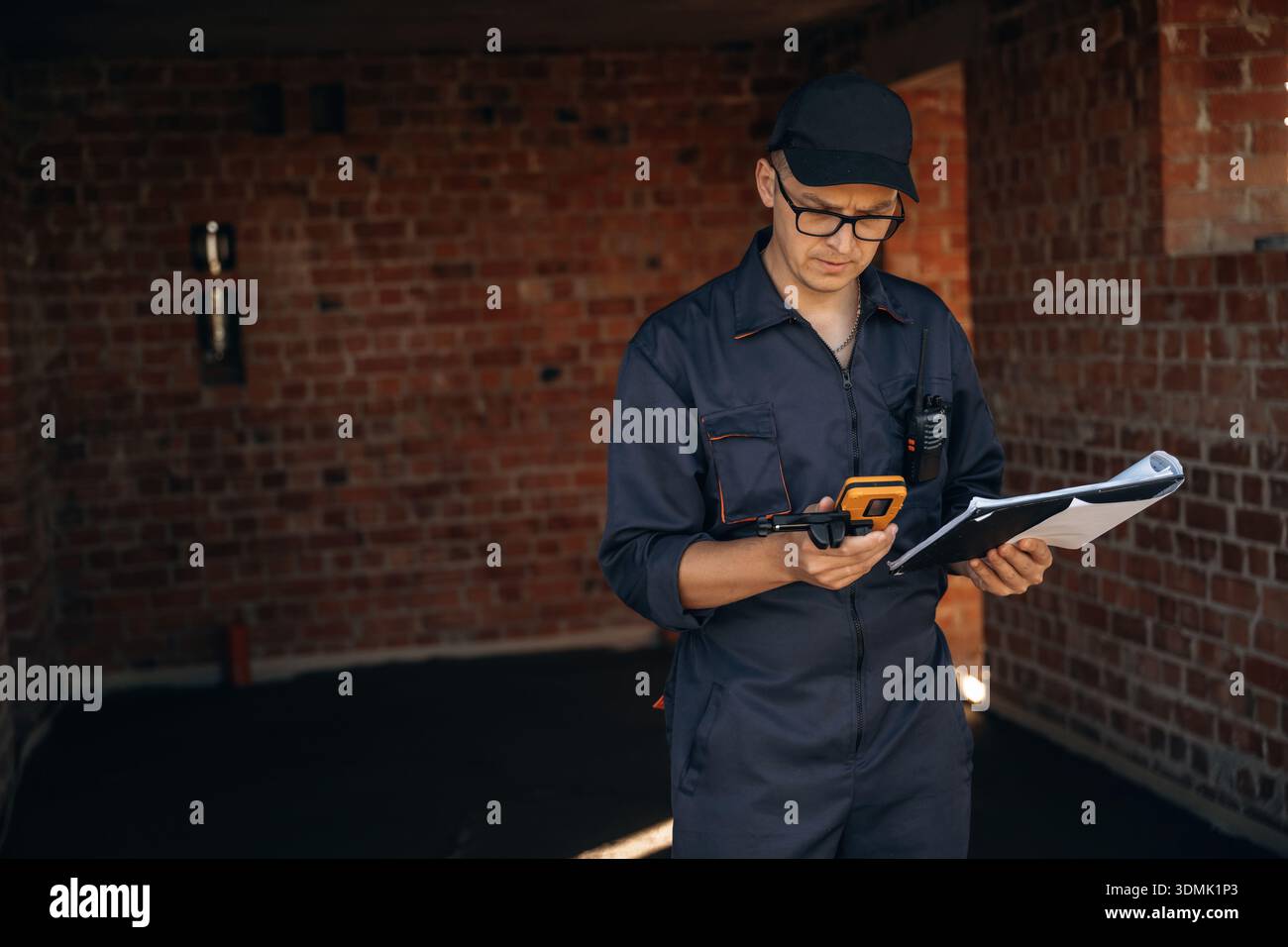 Documents and leveler tool are in hands. Builder is working on construction site, unfinished building. Stock Photo