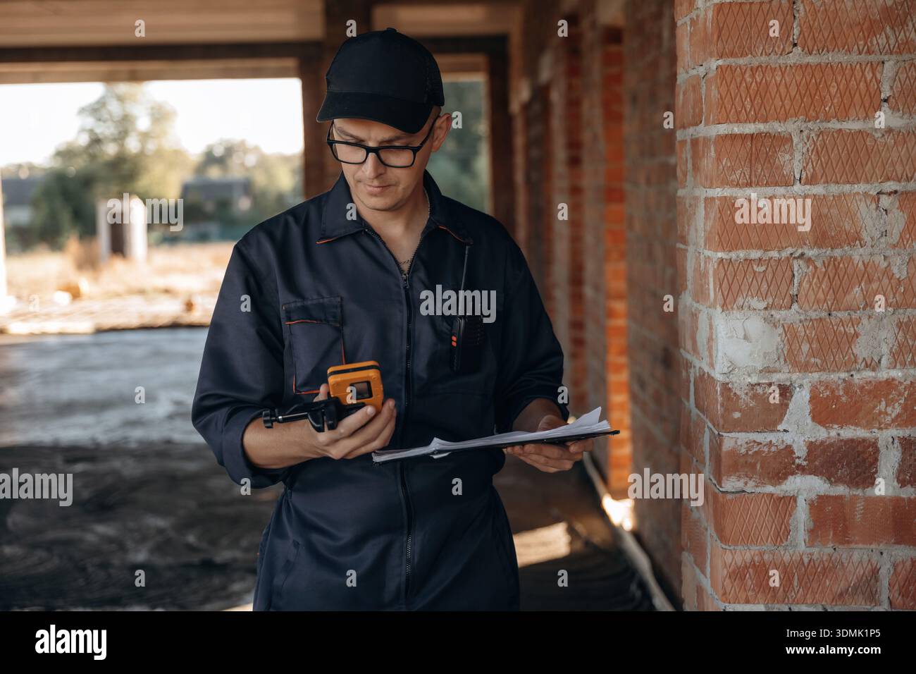 Documents and leveler tool are in hands. Builder is working on construction site, unfinished building. Stock Photo