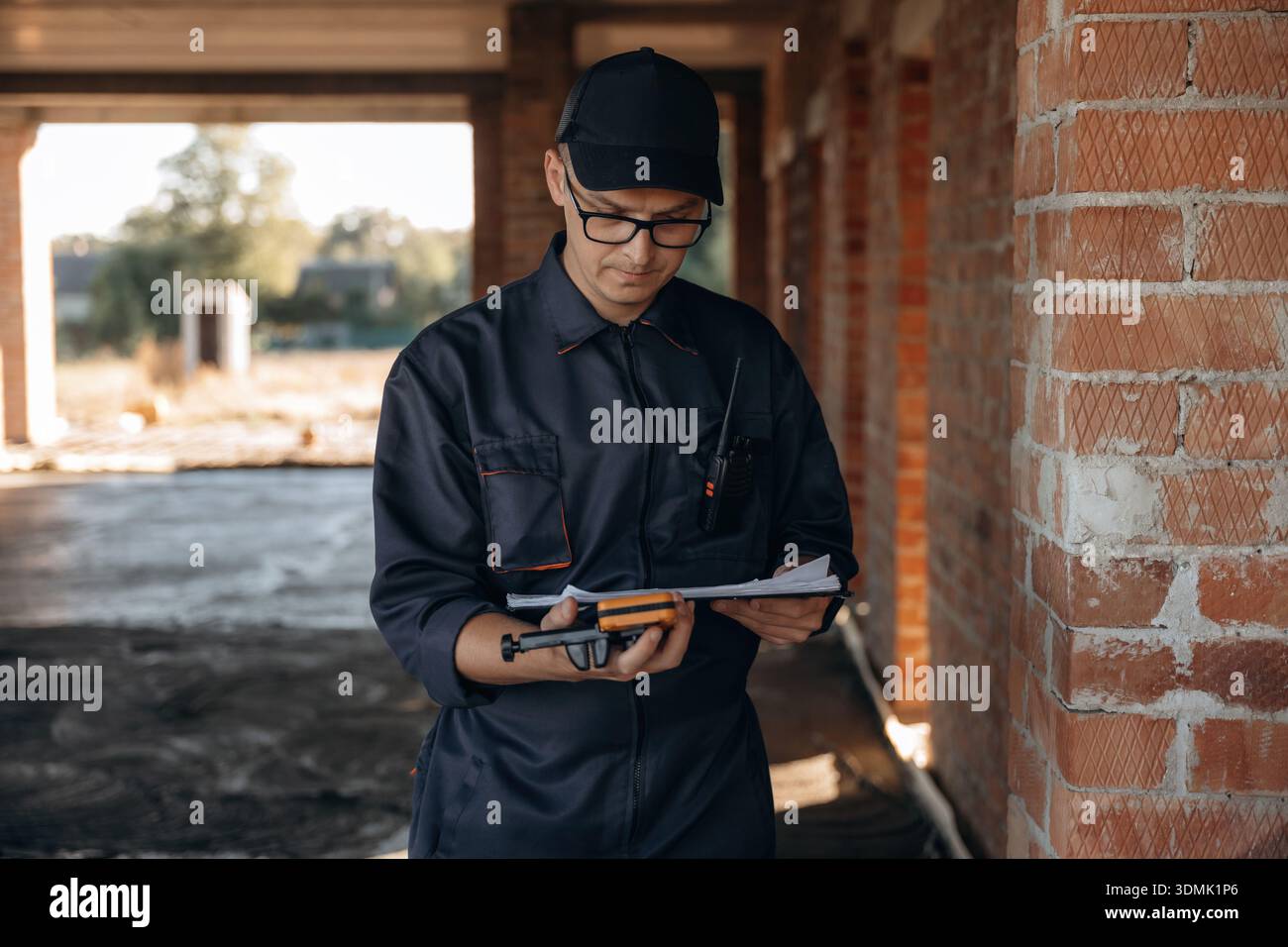 Documents and leveler tool are in hands. Builder is working on construction site, unfinished building. Stock Photo