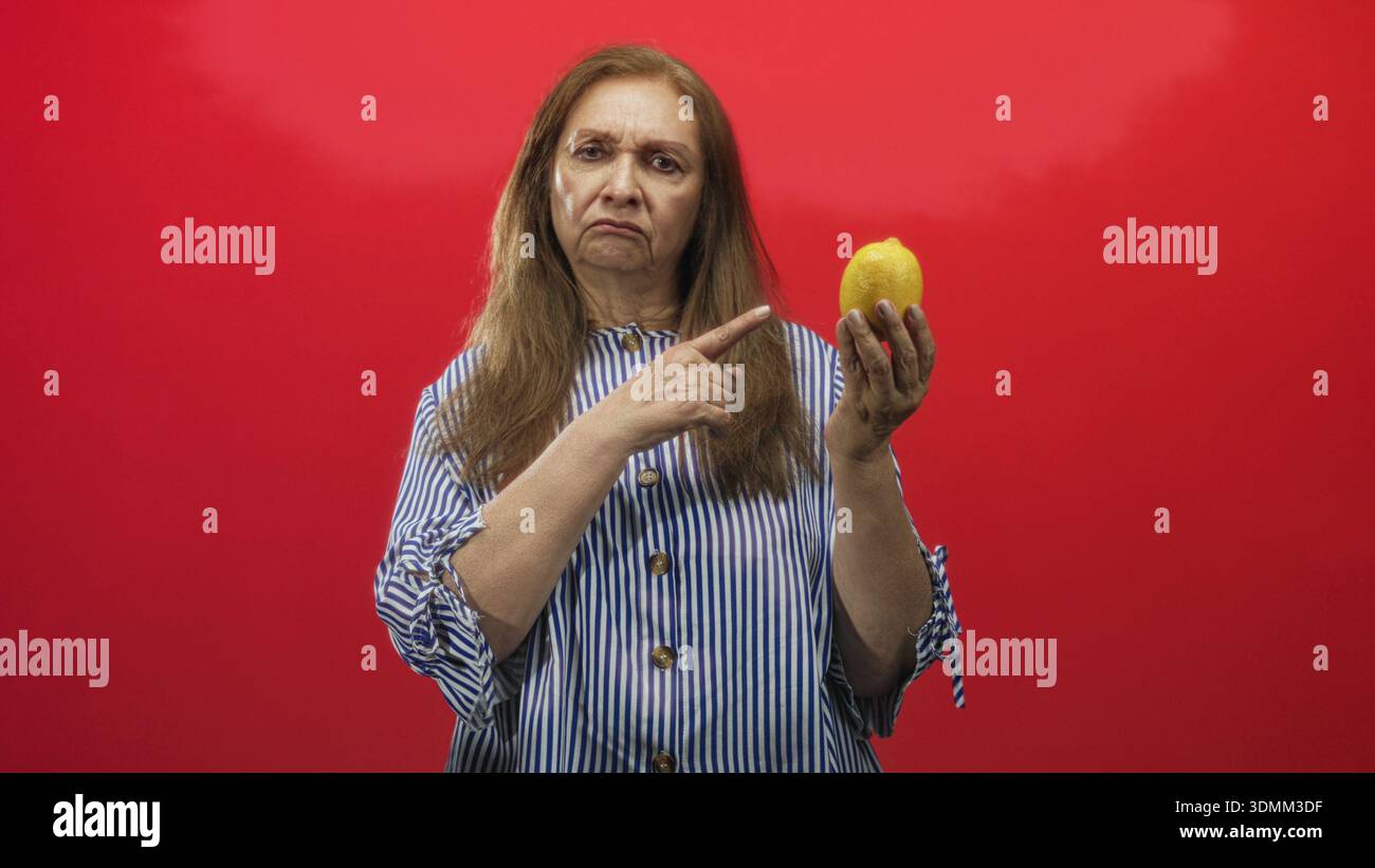 Senior hispanic woman holds lemon and points finger at lemon in studio wearing striped blouse; sourness warning disapproval. Stock Photo