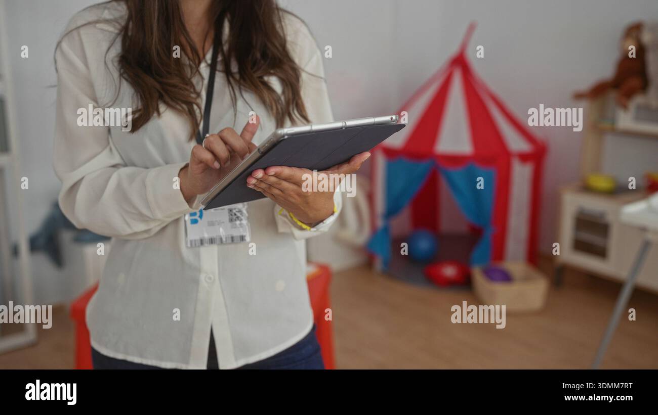 Hispanic woman using tablet in kindergarten classroom surrounded by colorful toys showcasing a preschool learning environment indoors. Stock Photo