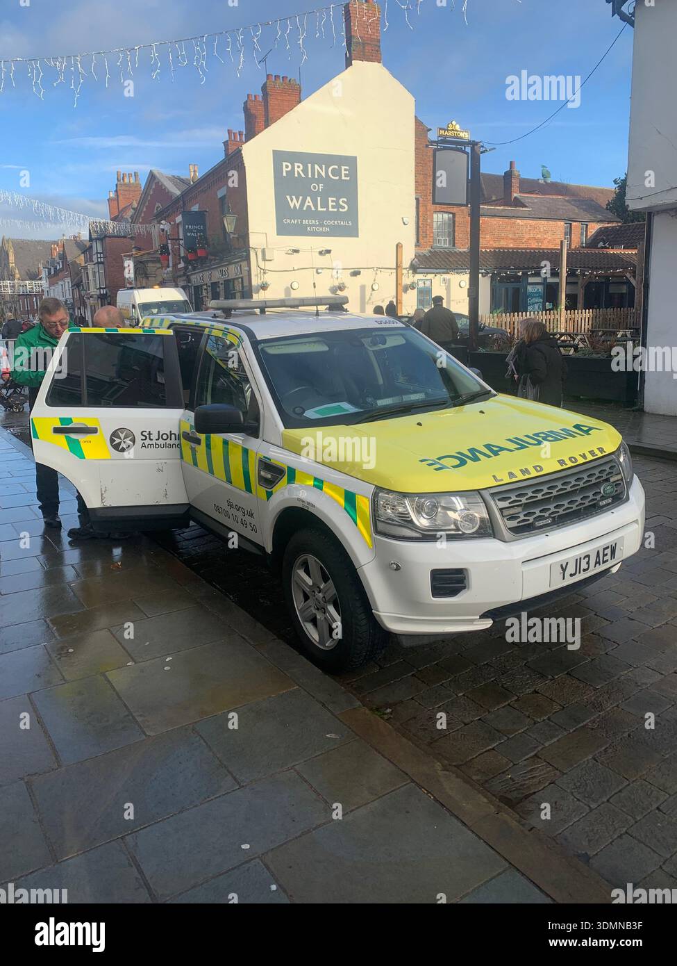 A St Johns Ambulance emergency response vehicle parked on a wet town centre street as crew members stands by the open rear door in Lincoln England - Smartphone Captured Stock Image