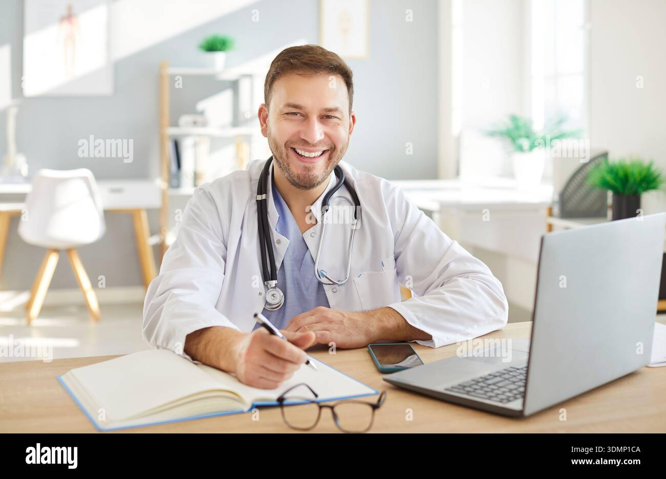 Happy smiling male doctor in white uniform working online on computer, learning, improving  Stock Photo