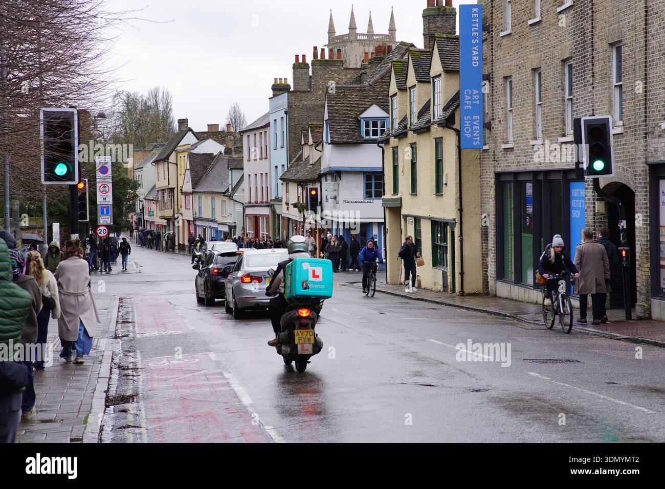 Delivery scooter rider on wet historic street in Cambridge city centre with colourful buildings and pedestrians. Cambridge,England Stock Photo
