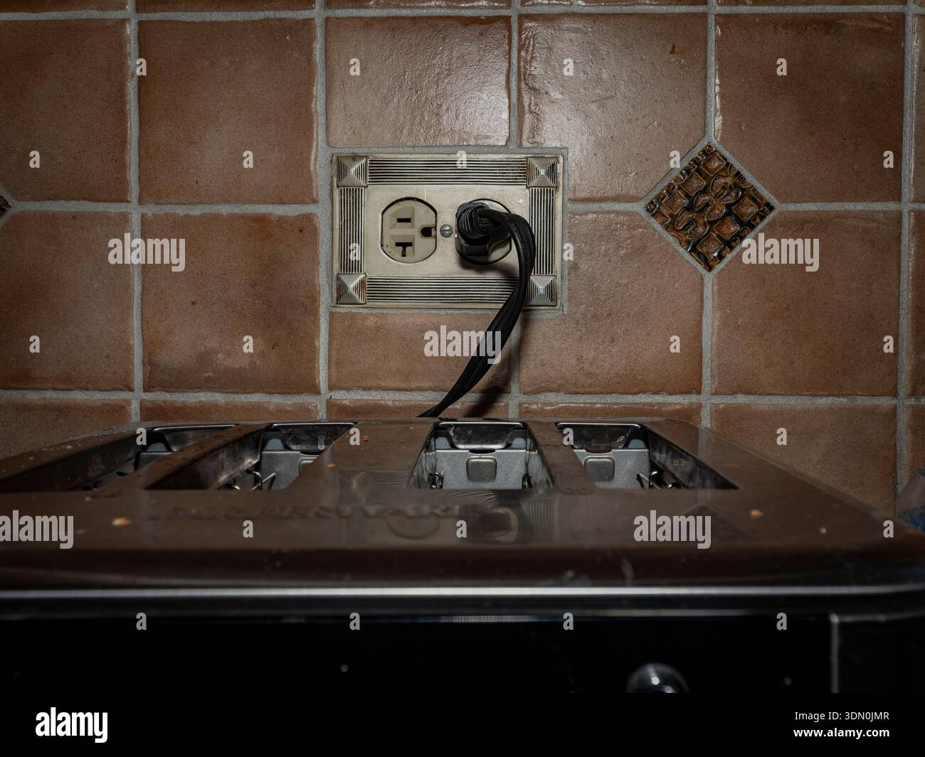 Toaster with power cord plugged into a wall outlet against brown tiled backsplash, captured in warm indoor lighting with household realism. Stock Photo