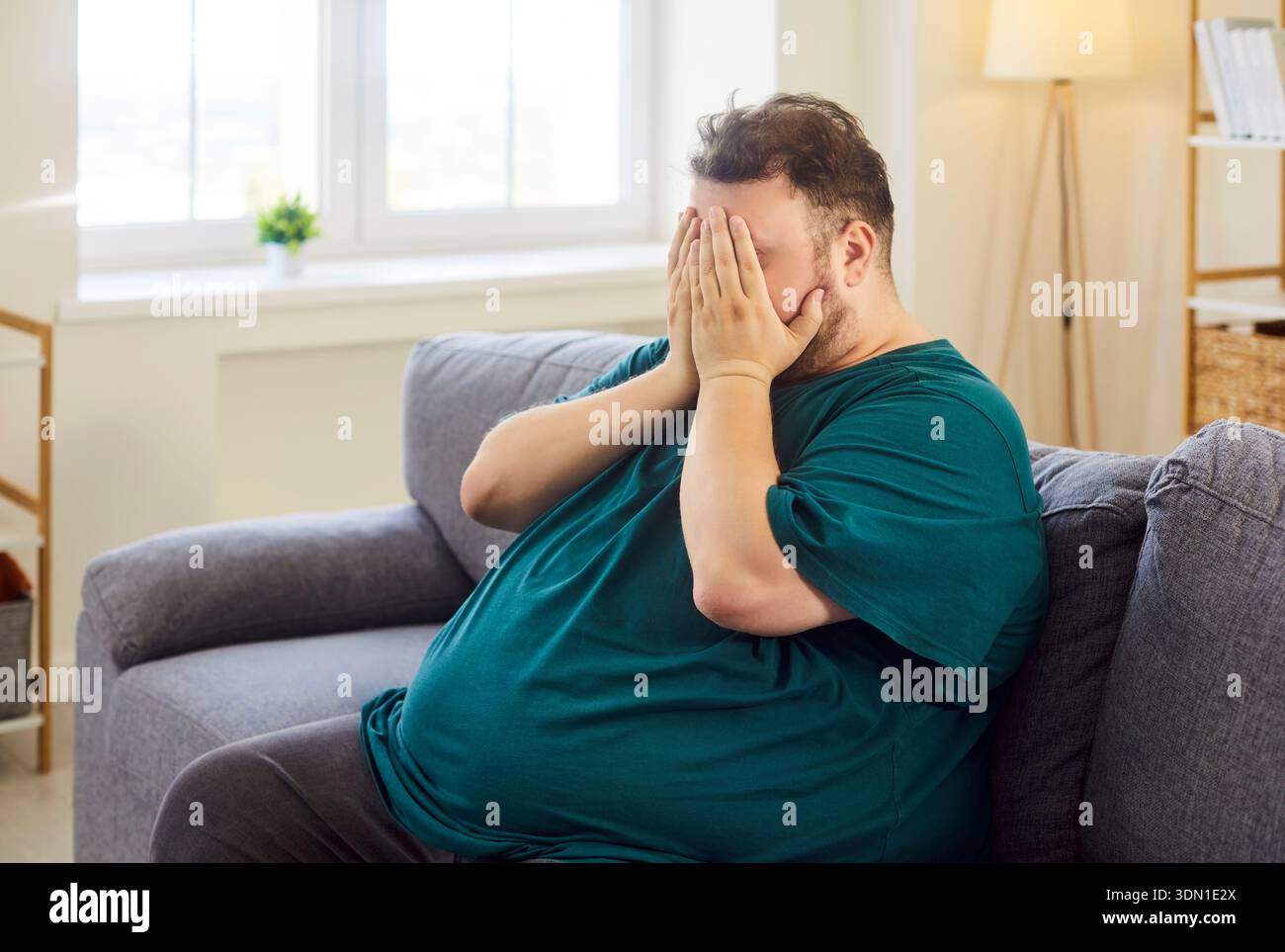 Anxious overweight man covering face on sofa at home, feeling stressed and tired Stock Photo