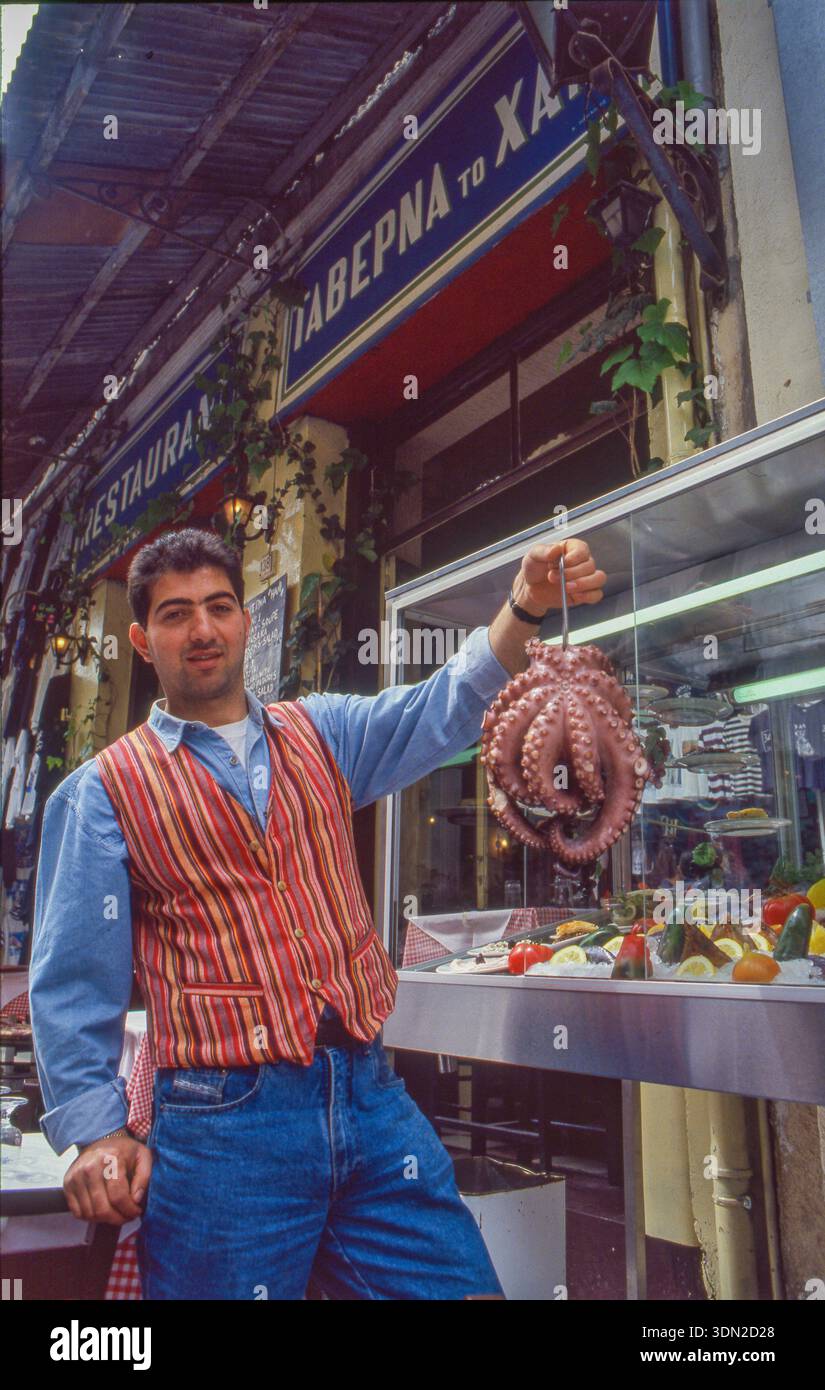 Greece, Athens - A waiter in a restaurant or tavern shows an octopus in front of a display case with seafood and vegetables. Stock Photo