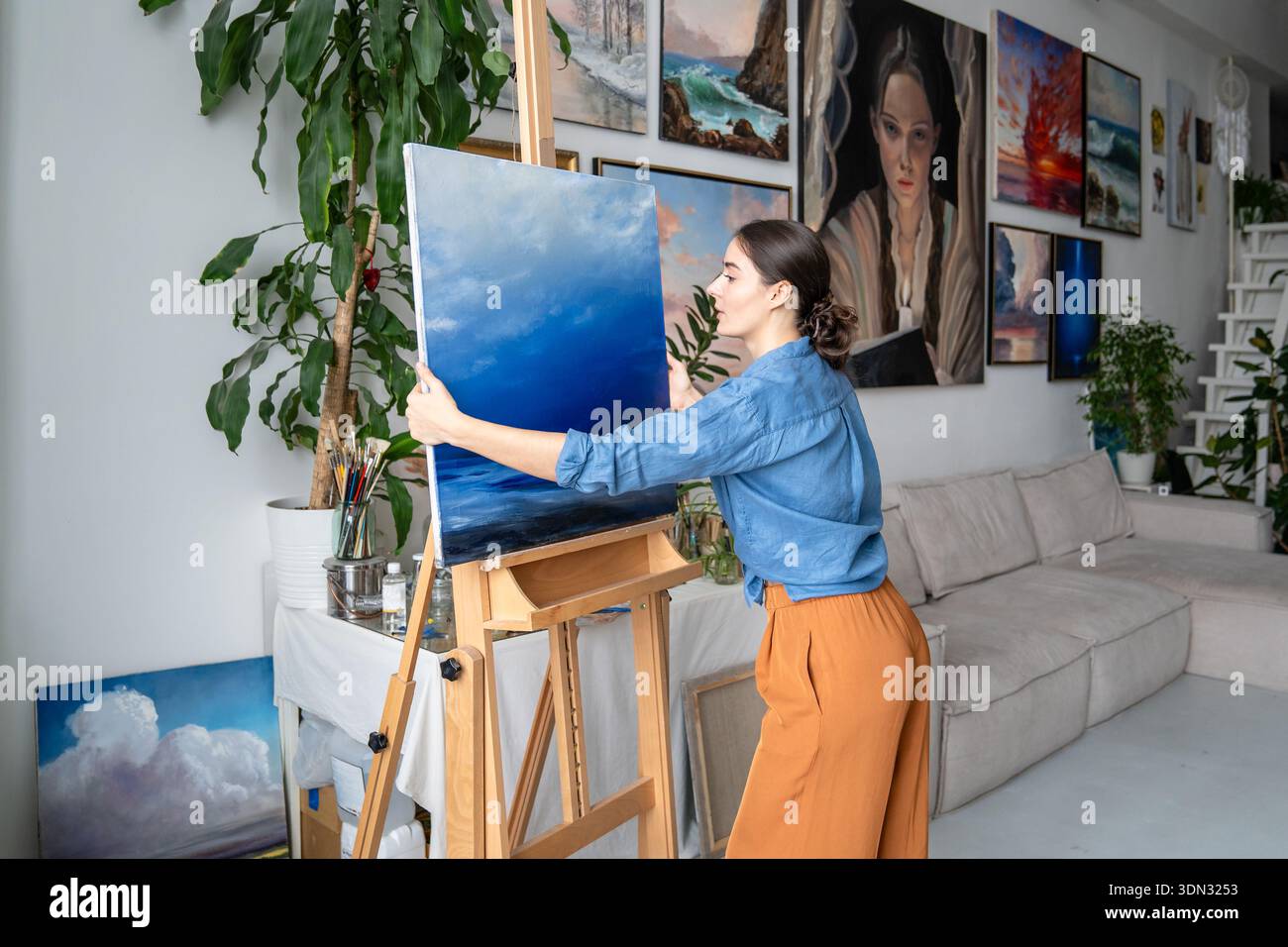 Focused woman artist working on painting in loft art gallery, adjusting finished artwork on easel. Stock Photo