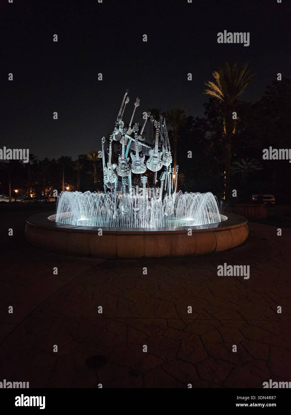 EDITORIAL USE ONLY-ORLANDO, FL-JANUARY 2026: A night view of the illuminated guitar fountain located at the entrance of the Hard Rock Hotel at Universal Orlando Resort. The circular stone fountain features a central sculpture of multiple electric guitars with water jets lit from below, surrounded by palm trees against a dark sky. - Smartphone Captured Stock Image