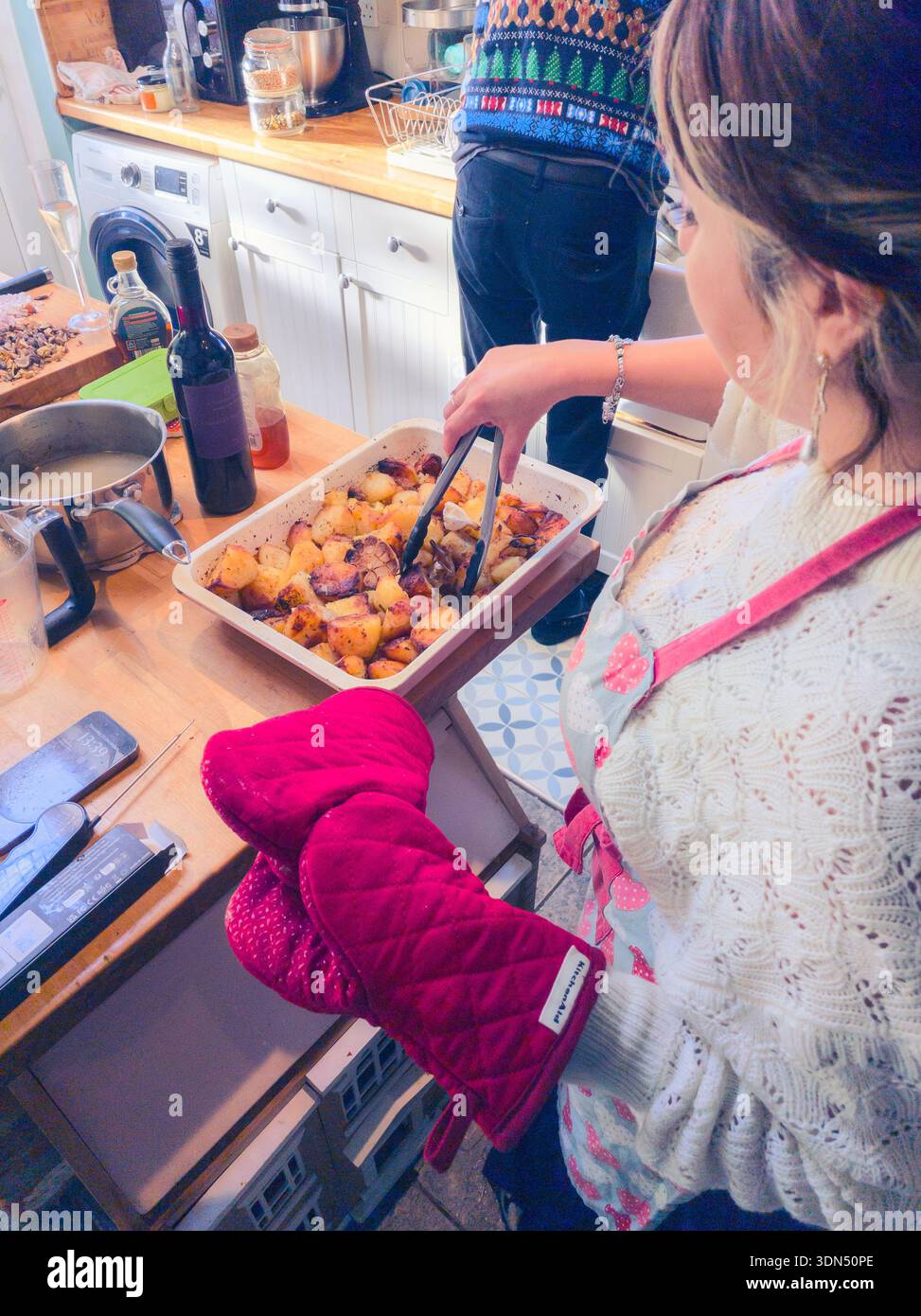 Millennial woman cooking roast potatoes for Christmas dinner wearing an oven glove and turning them with tongs & husband doing the washing up, UK - Smartphone Captured Stock Image