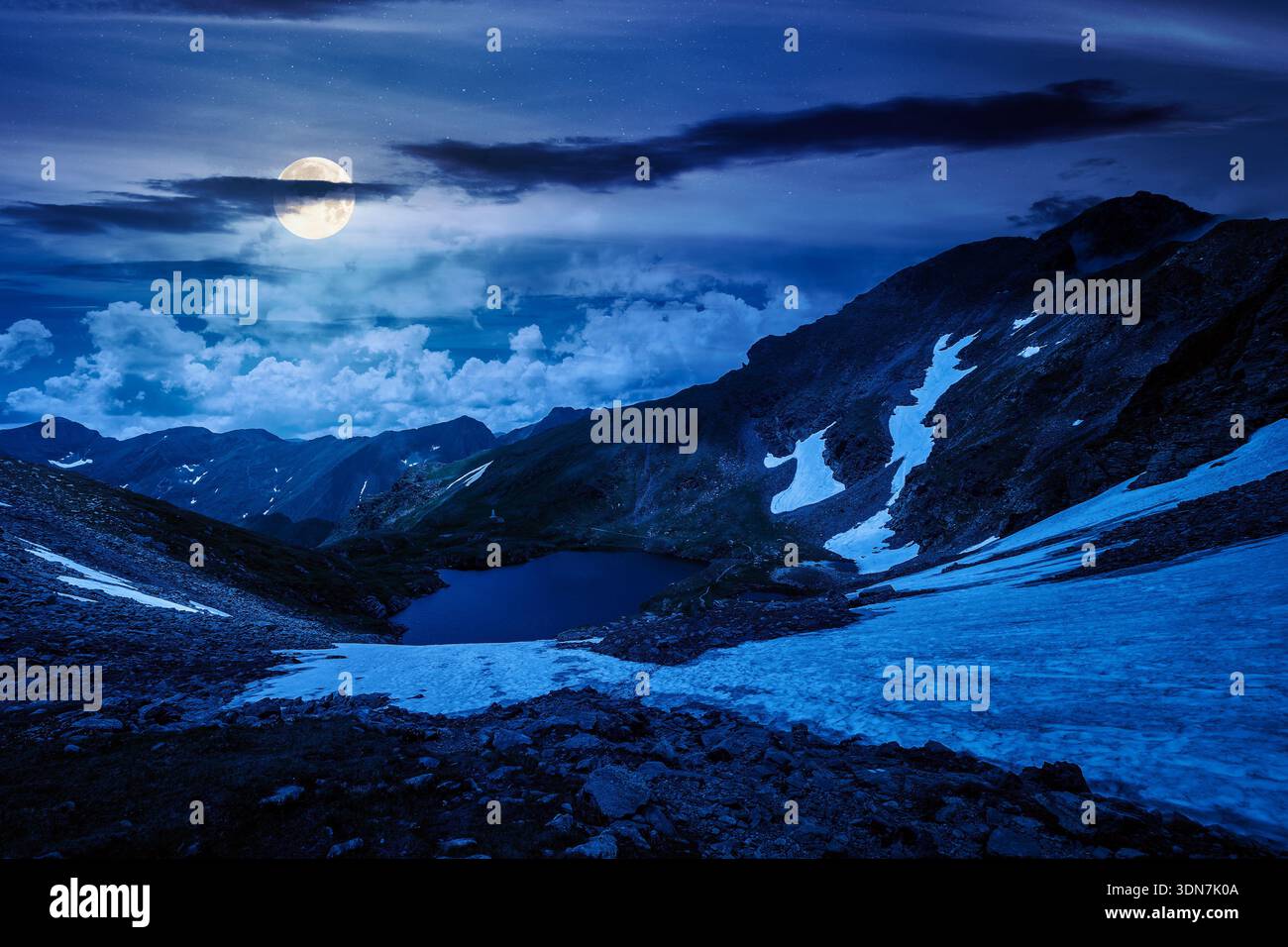 mountain lake in alpine summer landscape at night. beautiful view surrounded with clouds on a dark sky in full moon light. fresh water, snow and rocky Stock Photo