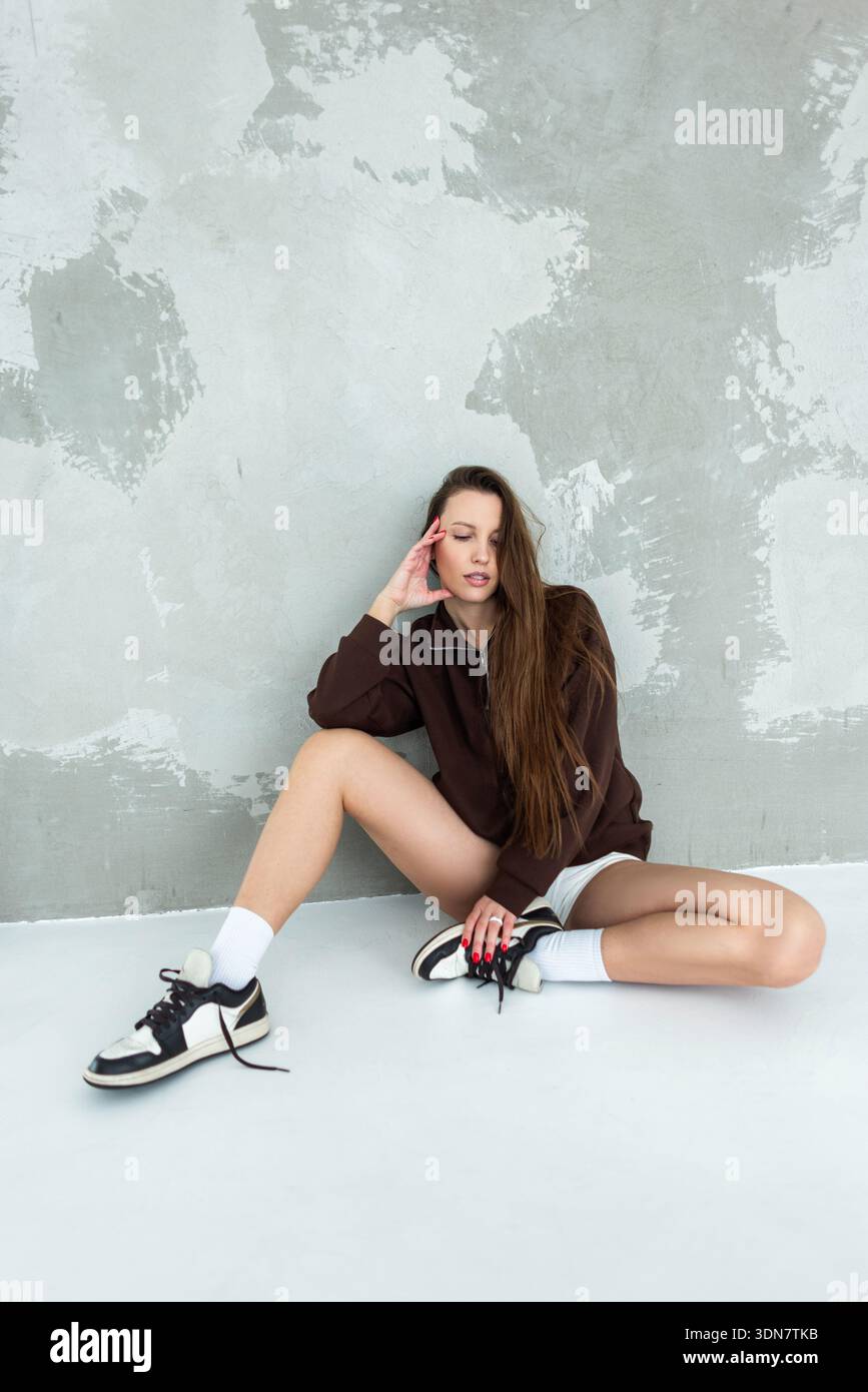 Slender woman posing in a trendy brown sweatshirt and white shorts. Sitting cross-legged on the floor against a textured plaster background. Natural l Stock Photo