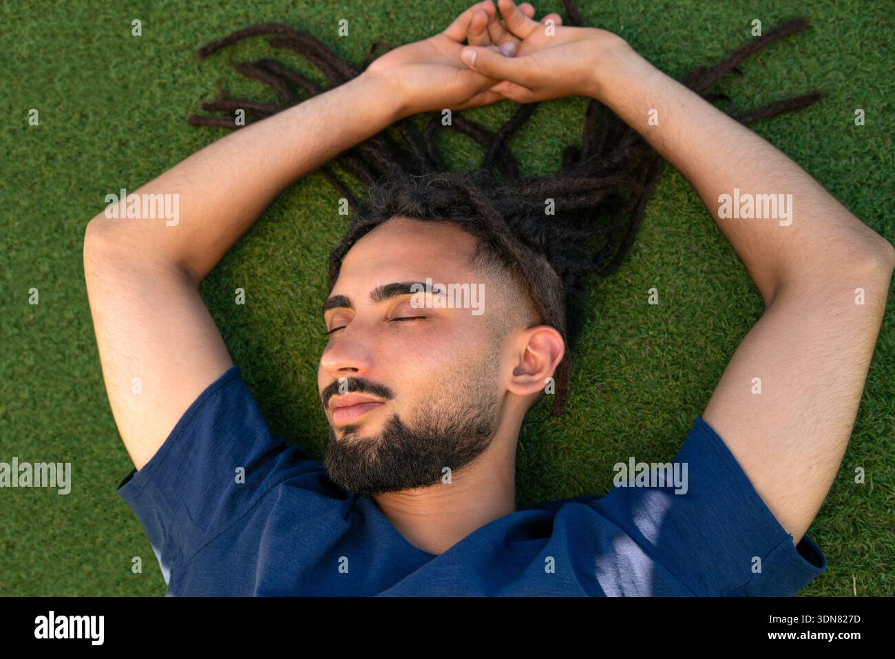 Young man with dreadlocks lying on green grass, feeling peaceful Stock Photo