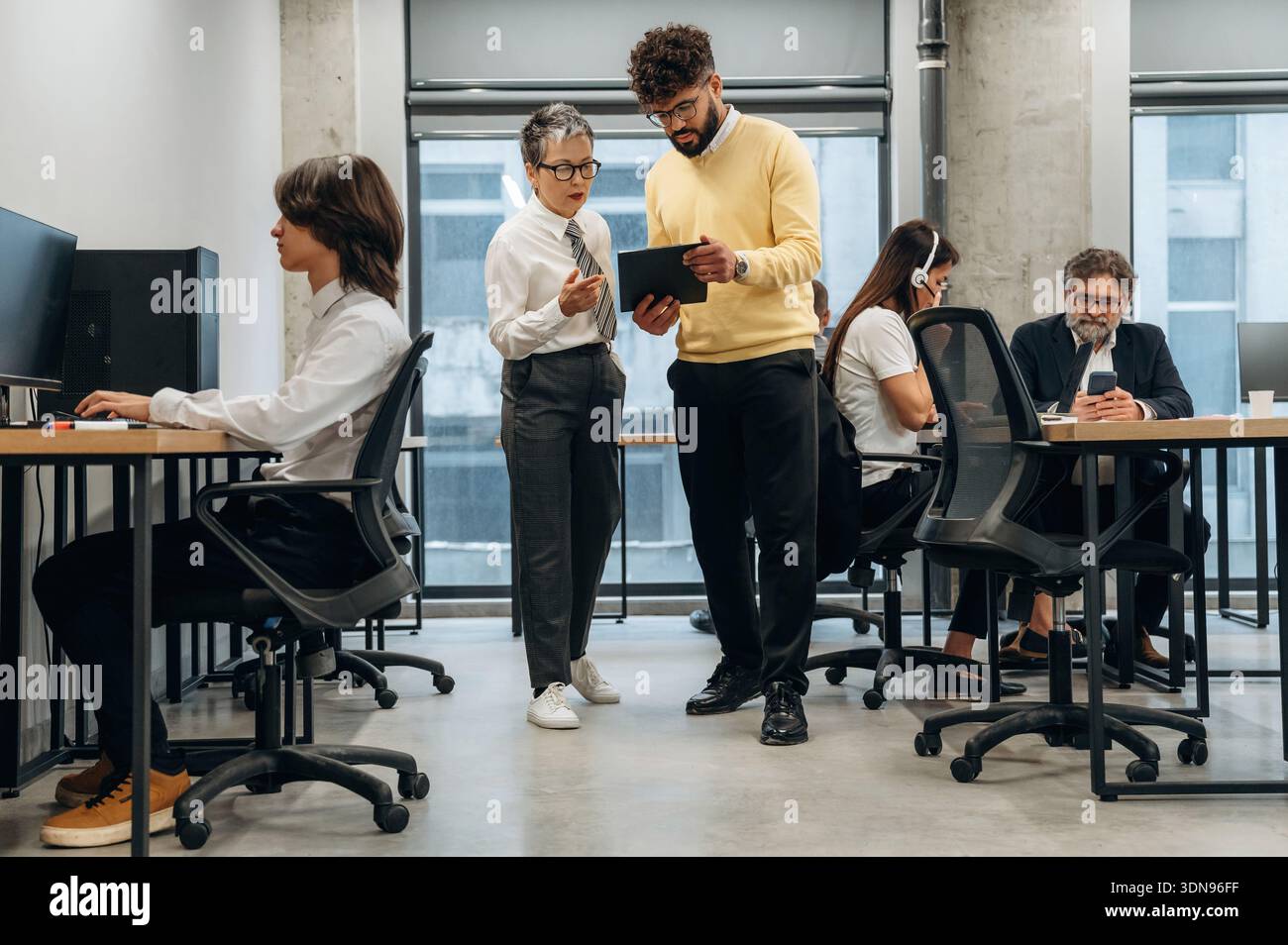 Two business professionals are collaborating in a modern office space, discussing project details while utilizing a digital tablet, surrounded by coll Stock Photo