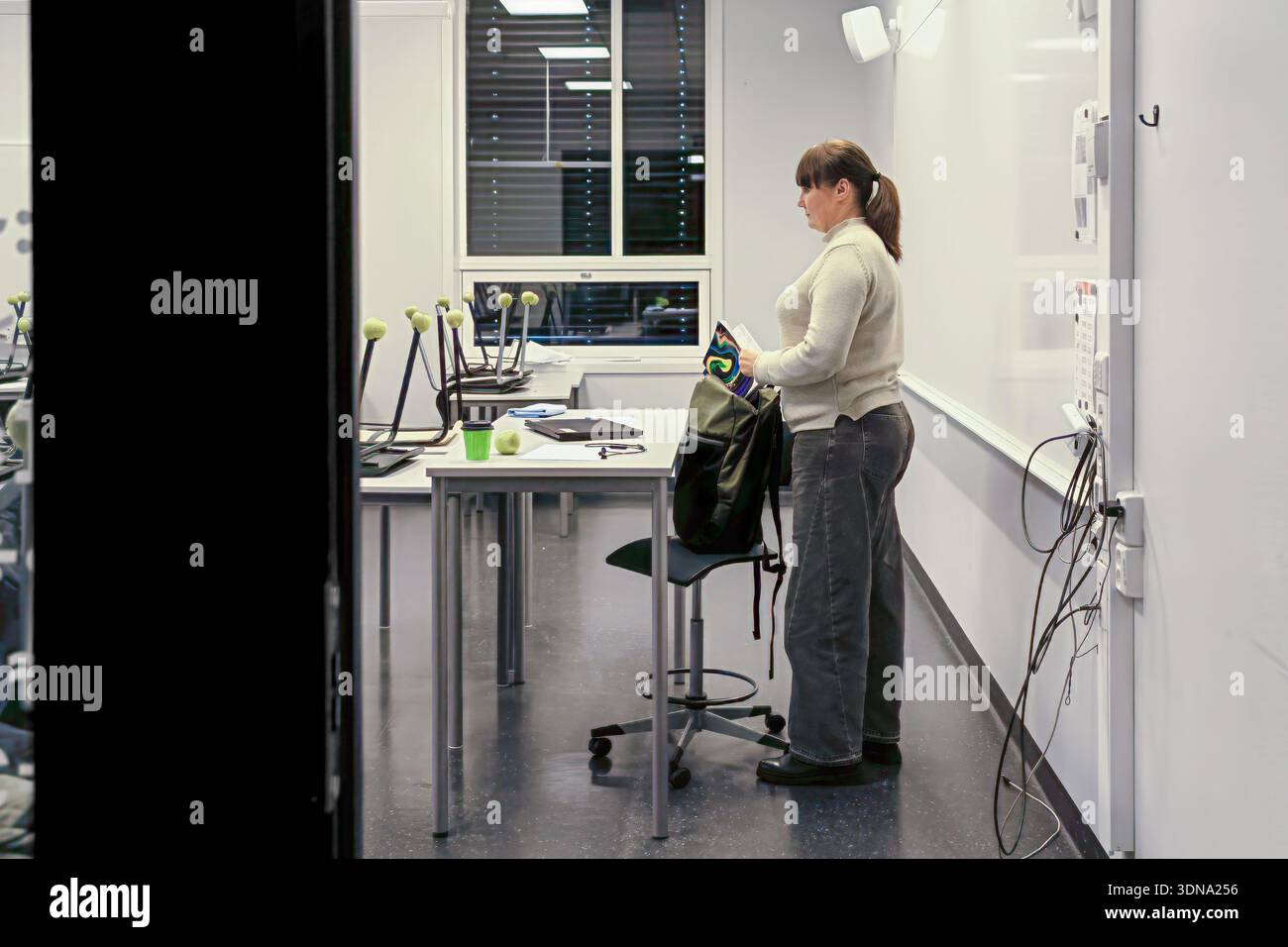Educator in a contemporary classroom, organizing materials on a desk, surrounded by empty chairs and large windows, fostering a positive educational a Stock Photo
