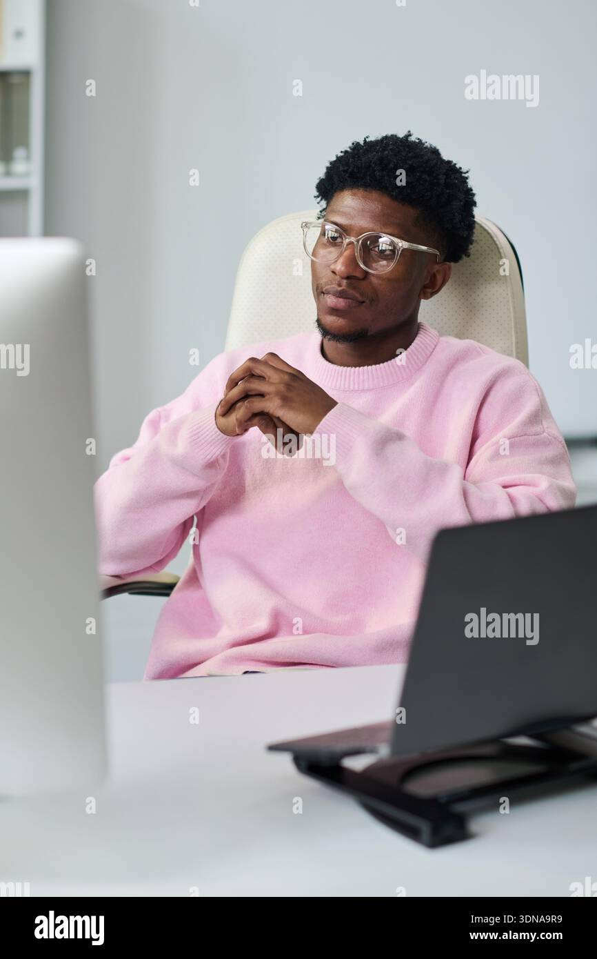 Young Black Man Sitting at Desk Using Computer and Looking Thoughtful Stock Photo