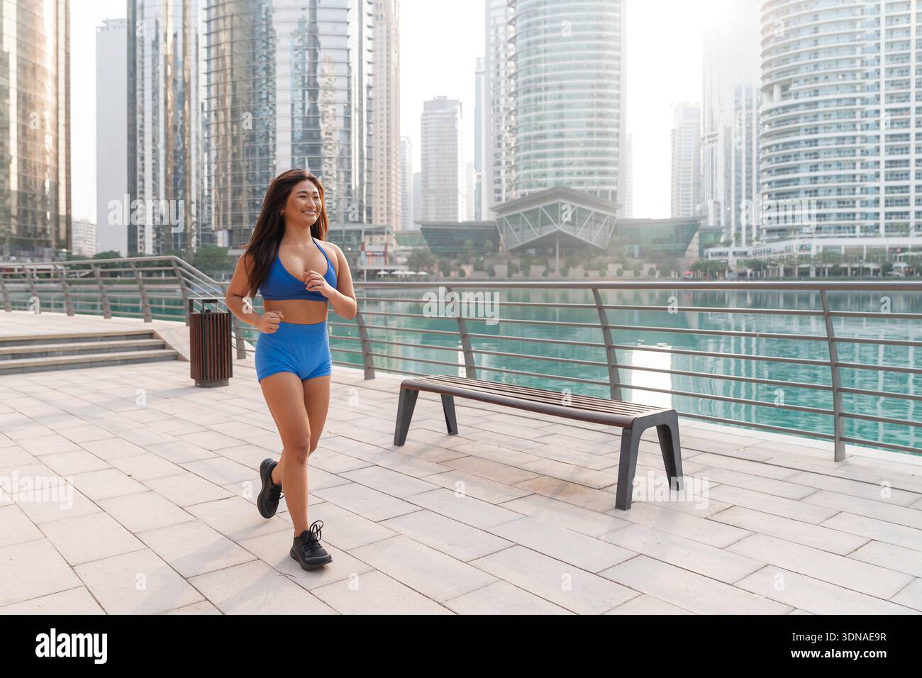 Asian woman jogging on urban promenade with city buildings and water behind her during morning light. Stock Photo