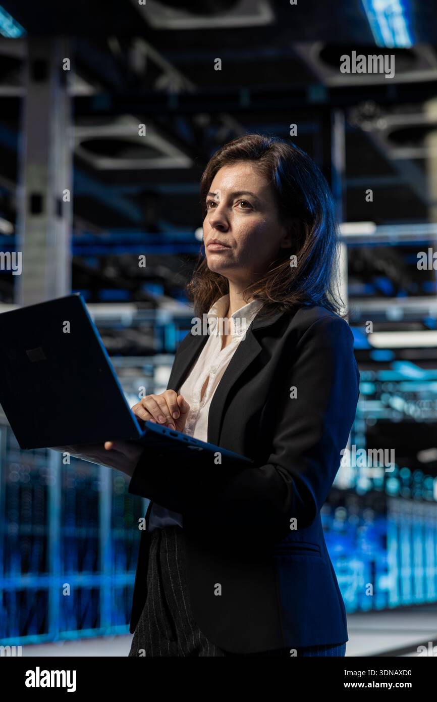 Data center female engineer using laptop, managing server virtualization. Computer scientist using notebook in server room, doing upgrades sustaining long term infrastructure reliability Stock Photo