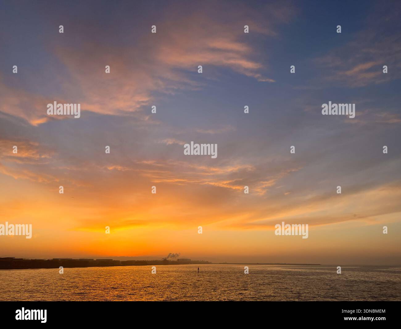 A blue sky with orange and yellow clouds hovers above a dark shoreline of a bay - Smartphone Captured Stock Image