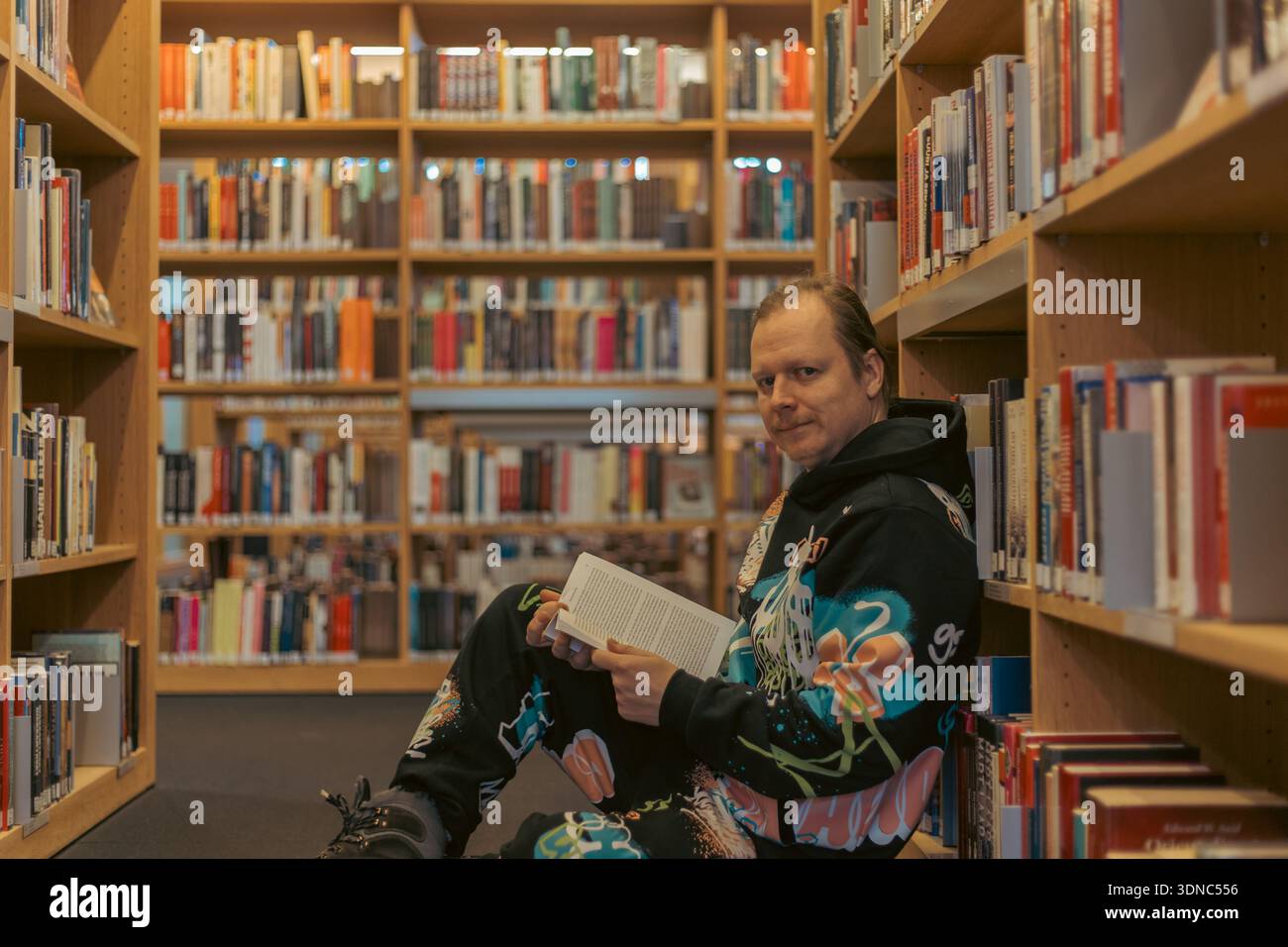 A young man sits on the floor in a well-lit, modern library, deeply engrossed in reading a book. Bookshelves filled with various books surround him, c Stock Photo