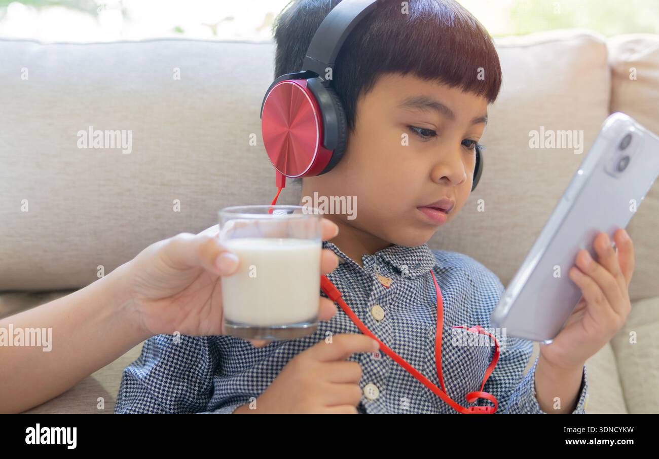 Adorable Asian baby boy with addiction phone and refusing his mouth facing away from milk from his mother. Kid's negative behavior. Stock Photo