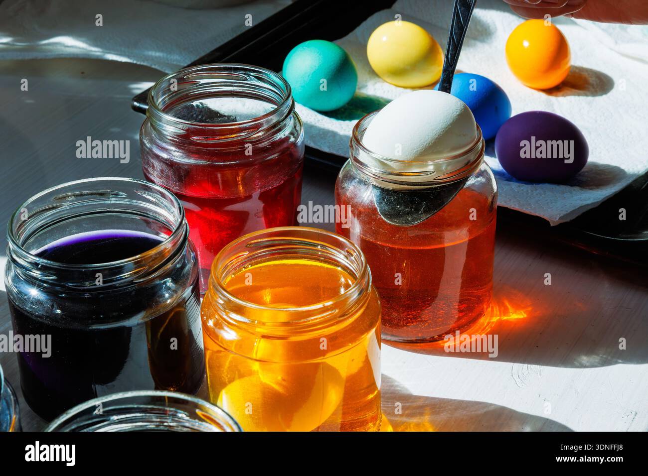 Spoon putting white egg into an orange dye jar with colorful dyed easter eggs nearby Stock Photo