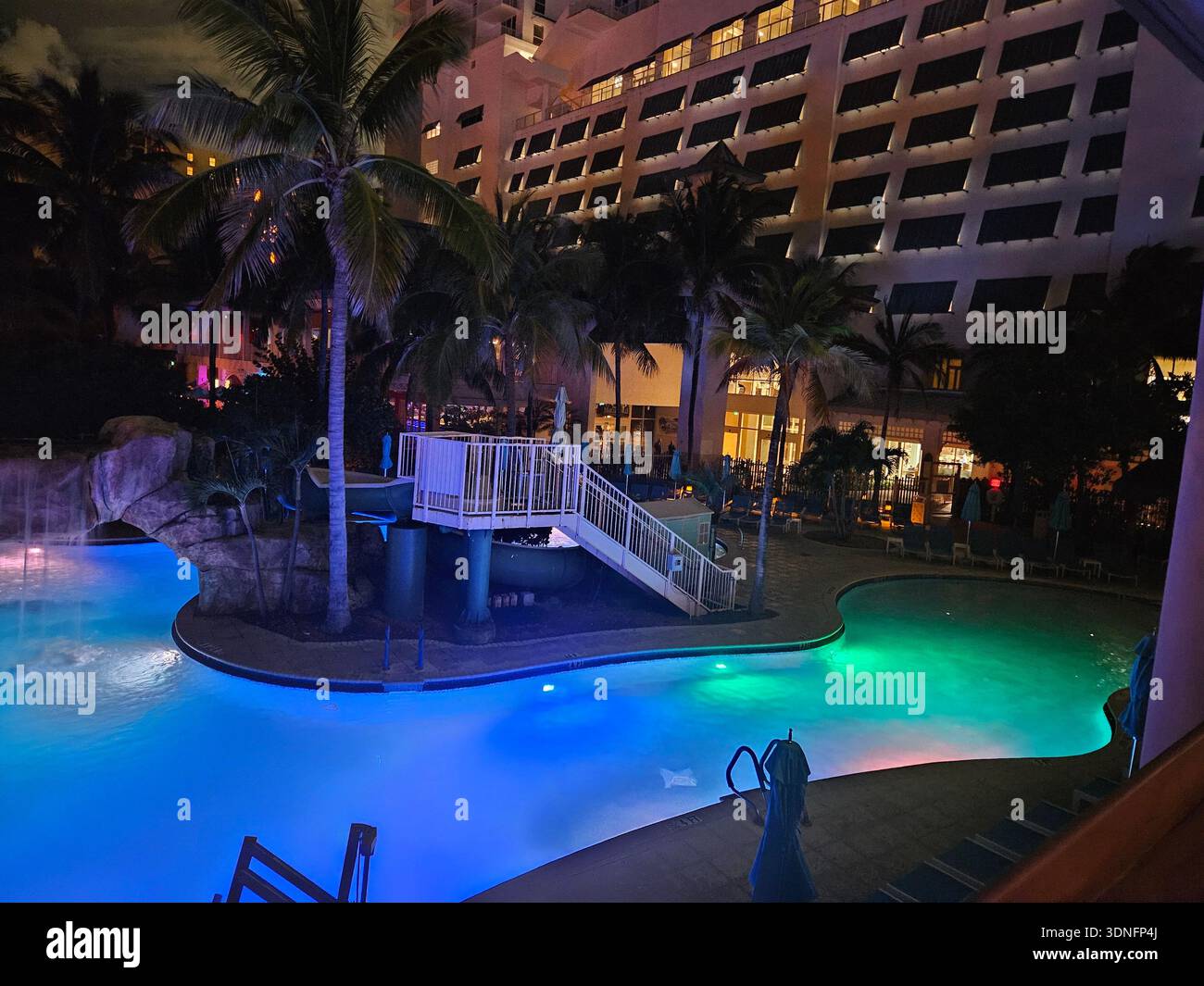 EDITORIAL USE ONLY- MARGARITAVILLE RESORT HOLLYWOOD BEACH FLORIDA: An evening view of the illuminated lagoon-style pool at the Margaritaville Hollywood Beach Resort. The scene features glowing blue and green underwater lighting, a rock waterfall, and a water slide, set against the backdrop of the resort’s tiered architectural balconies and tropical palm trees. This amenity-rich area is a centerpiece of the 349-room destination located on the Hollywood Beach Broadwalk. - Smartphone Captured Stock Image
