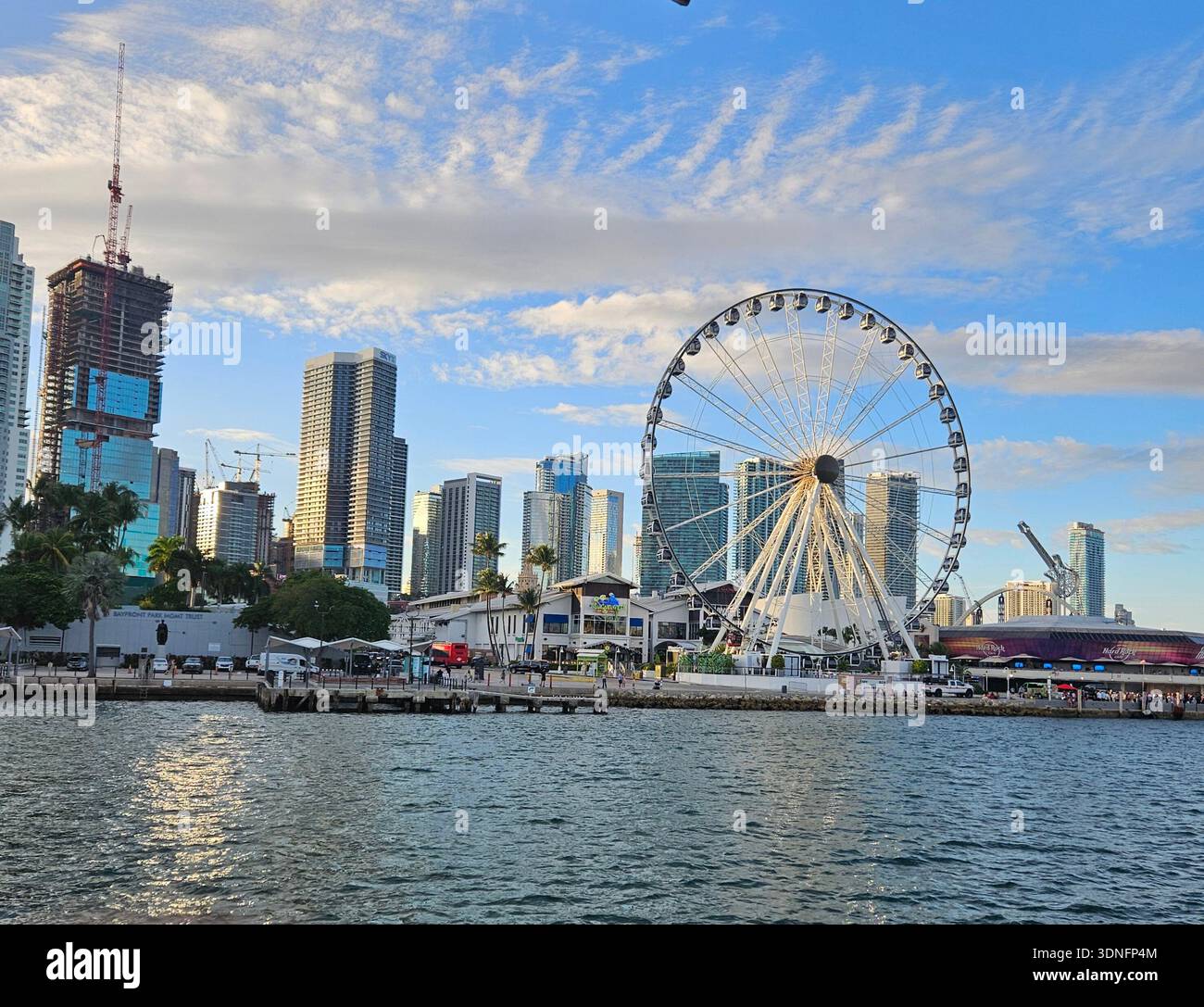 EDITORIAL USE ONLY- MIAMI FLORIDA: The Skyviews Miami observation wheel stands over Bayside Marketplace along the Biscayne Bay waterfront in Downtown Miami. The 176-foot tall Ferris wheel is pictured during the afternoon against a backdrop of city skyscrapers and the modern urban skyline. The scene captures the high-density development of the Brickell and Downtown districts, along with ongoing construction and maritime activity in the harbor. - Smartphone Captured Stock Image