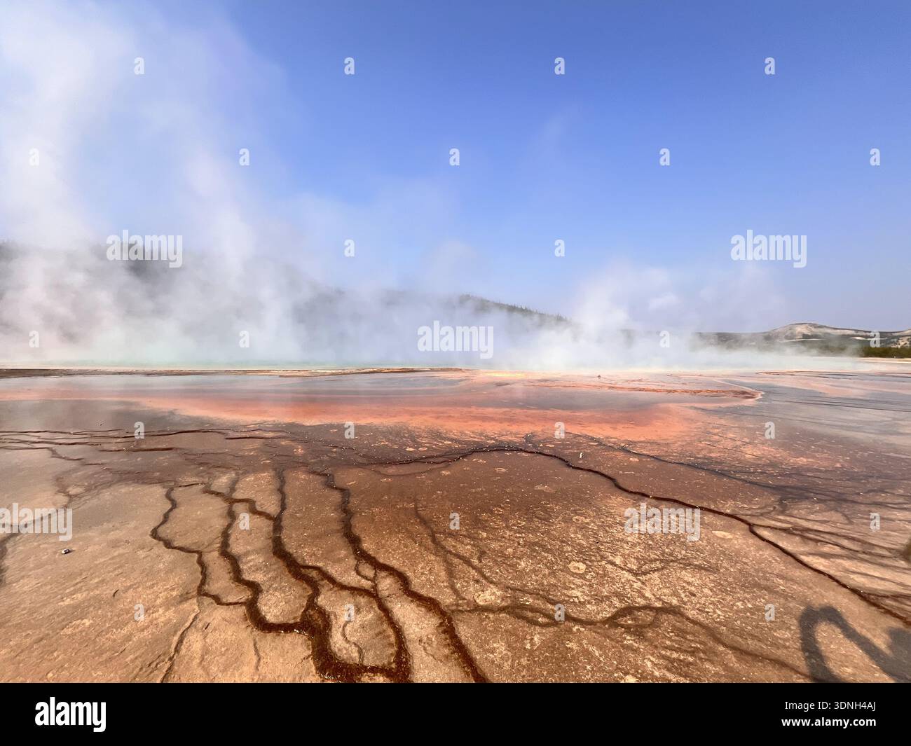 Steam rises from vivid orange and red mineral deposits in a geothermal landscape, creating a dramatic and colorful scene framed by hills and clear sky - Smartphone Captured Stock Image