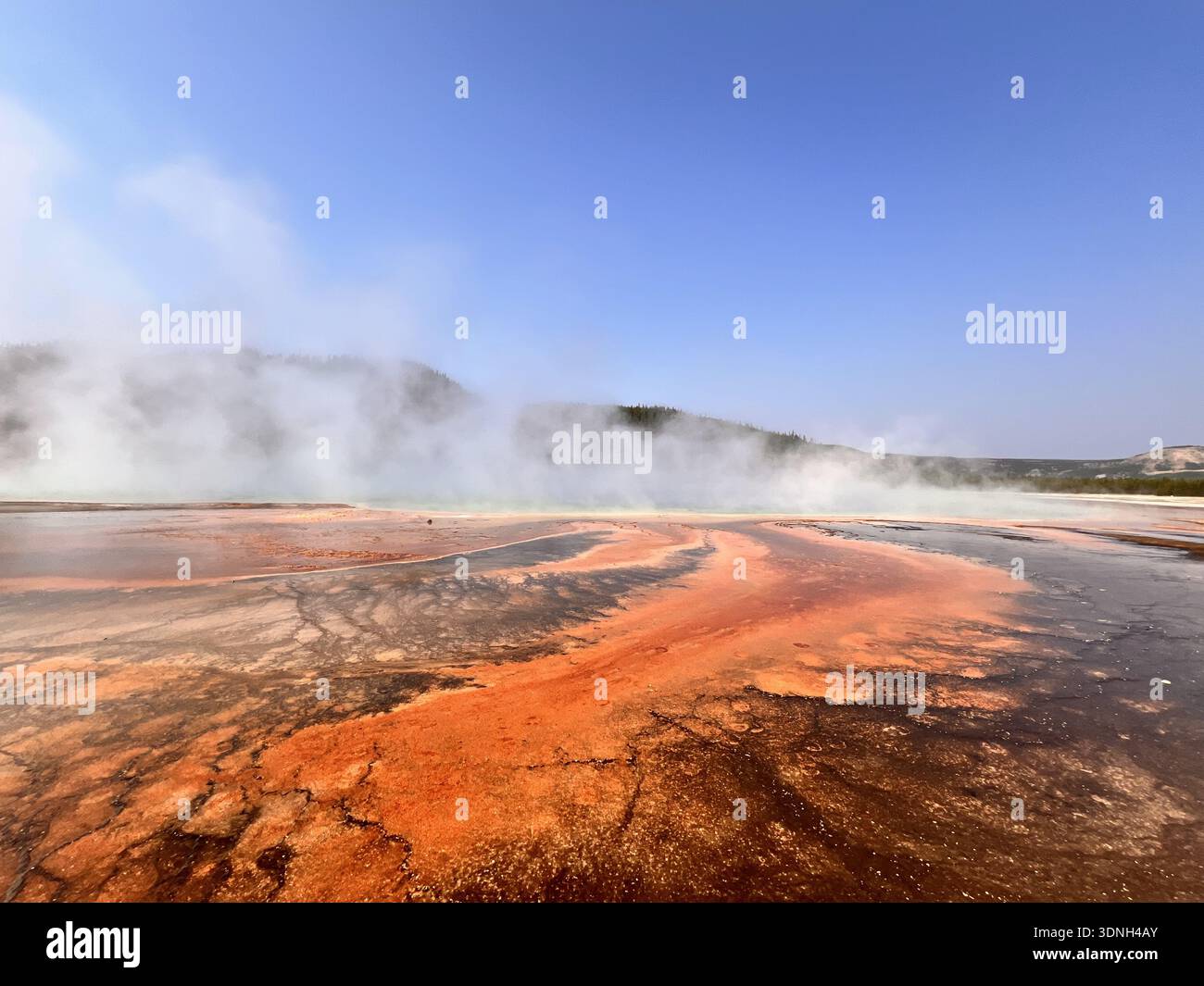 Steam rises from vivid orange and red mineral deposits in a geothermal landscape, creating a dramatic and colorful scene framed by hills and clear sky - Smartphone Captured Stock Image