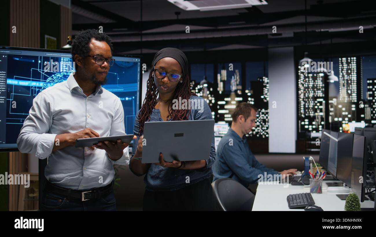 Tech agency colleagues collaborating to optimize AI performance in office late at night. Software developers use machine learning on devices for debugging, analyzing code Stock Photo