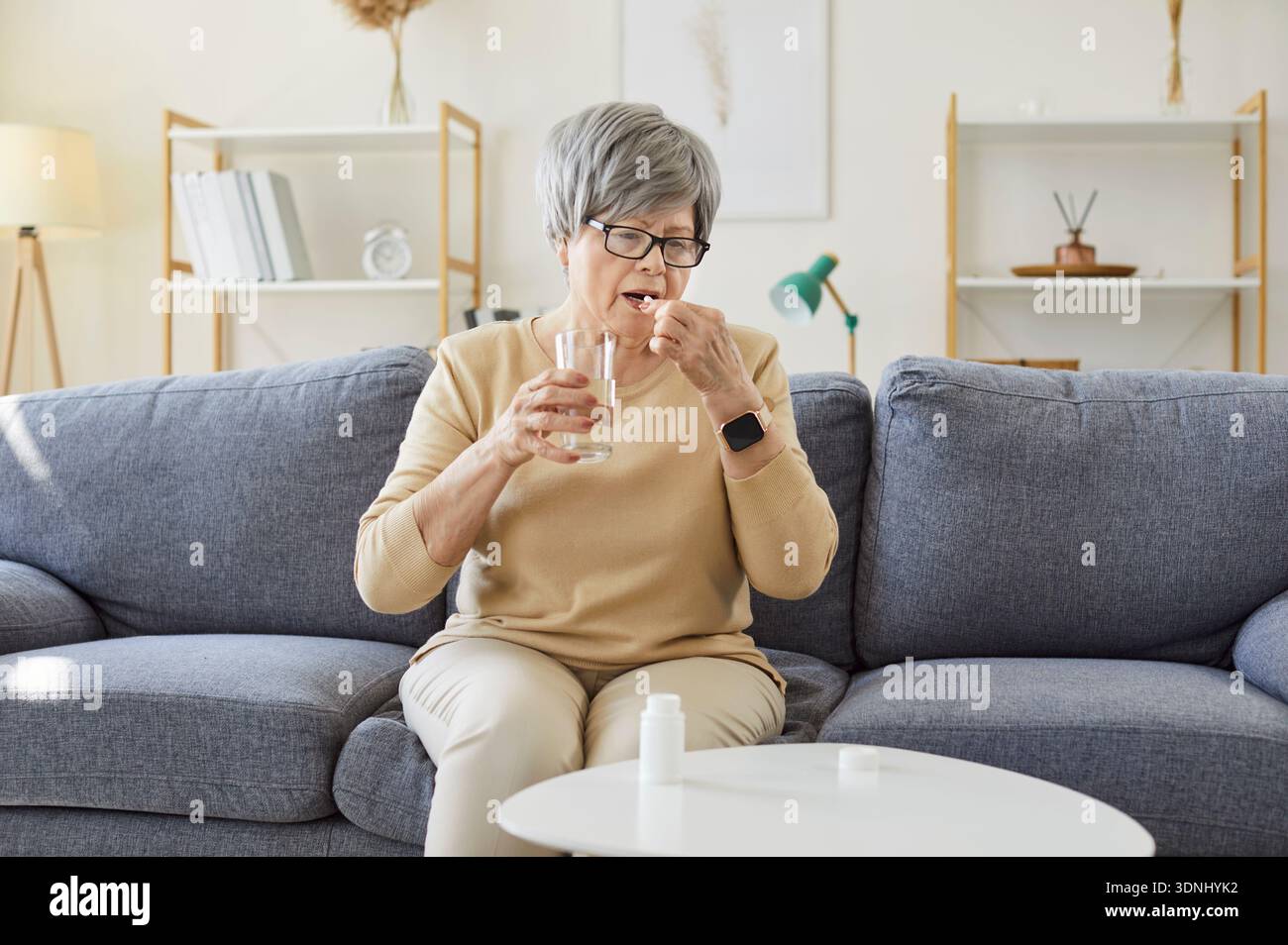 Senior woman taking pill dose at home, holding glass of water and capsule Stock Photo