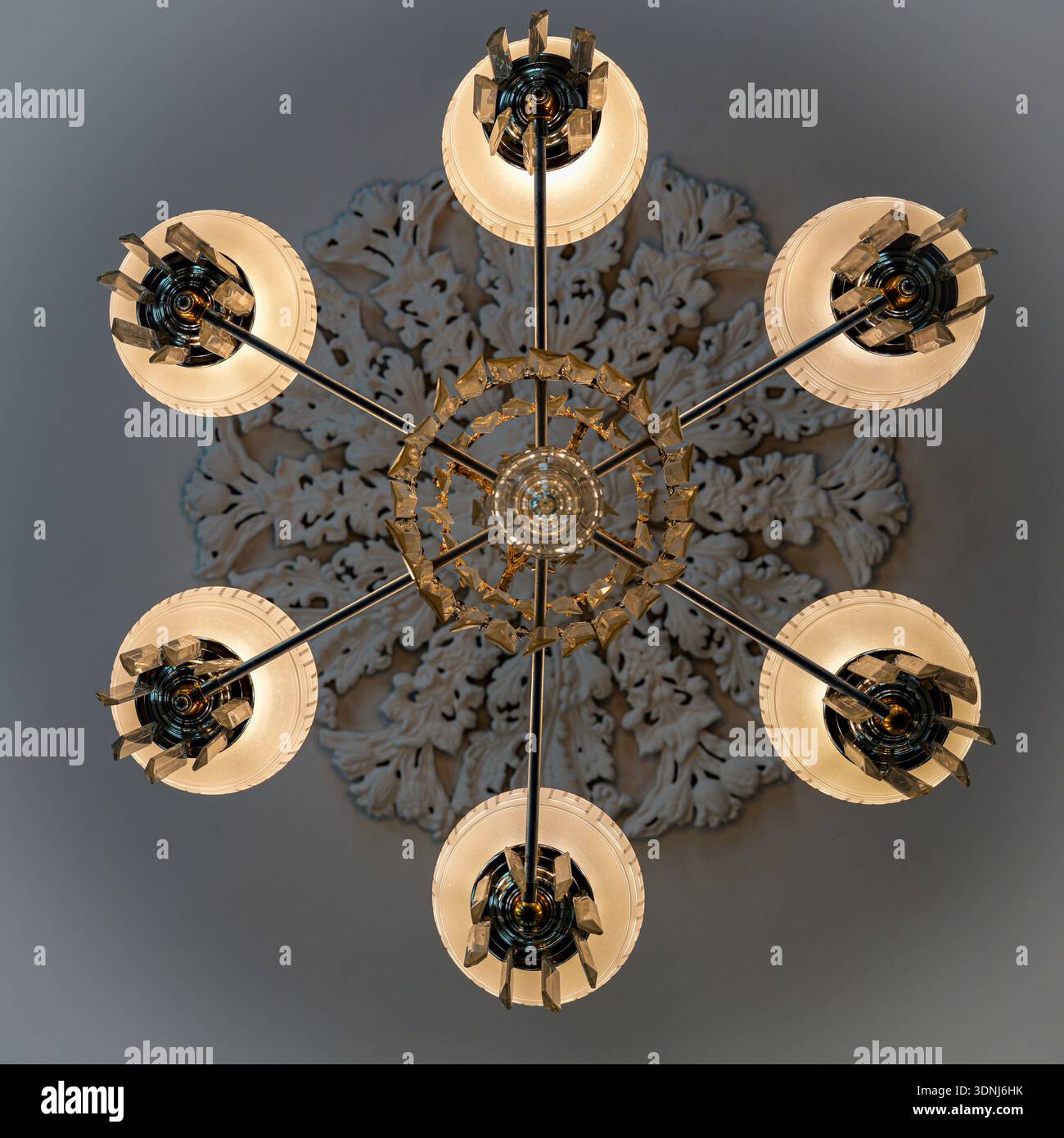 Looking straight up at a six light chandelier with glass accents radiating from an ornate ceiling medallion in a historic building interior. Stock Photo