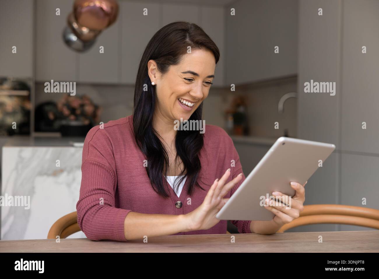 Woman using tablet while sitting at table in modern kitchen Stock Photo