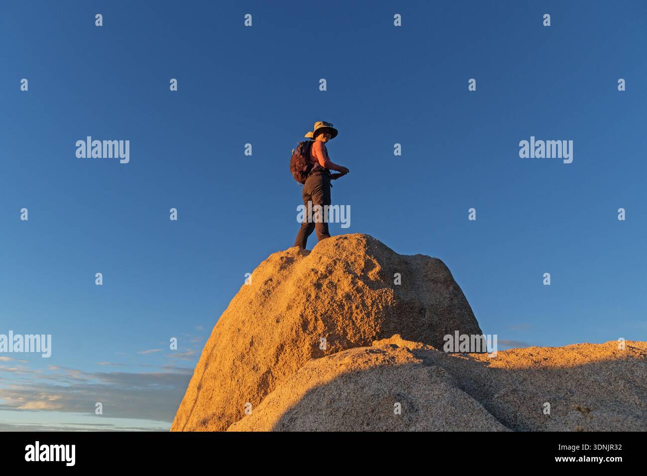 woman hiker with hat turned to the side on top of a boulder at sunset Stock Photo