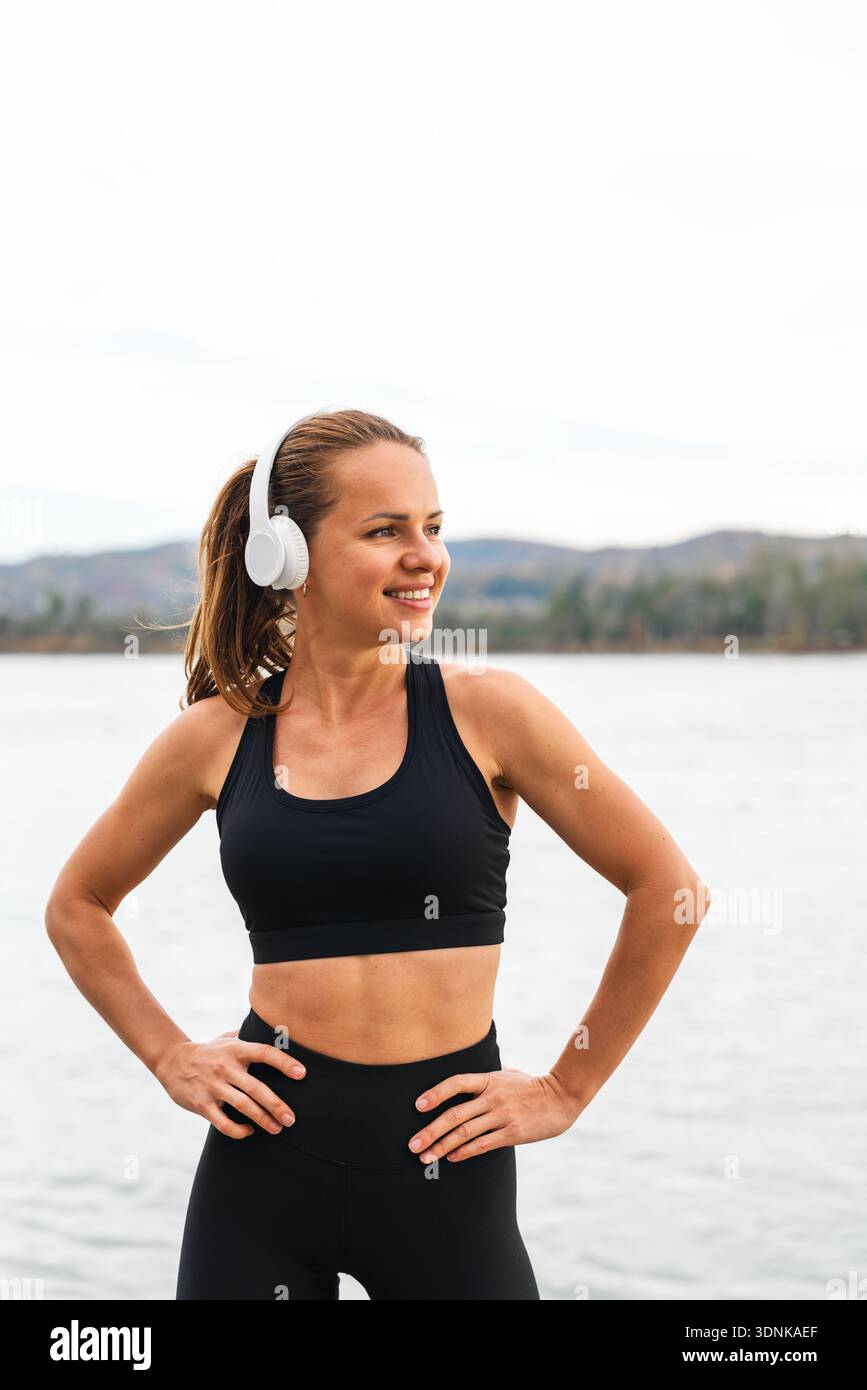 Smiling sporty female resting after workout and enjoying music in nature with river on background. Stock Photo