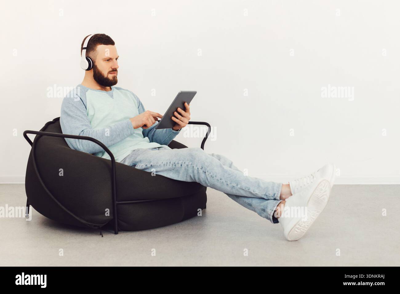 Serious Guy Using Tablet Wearing Earphones Sitting Indoor, Side View Stock Photo