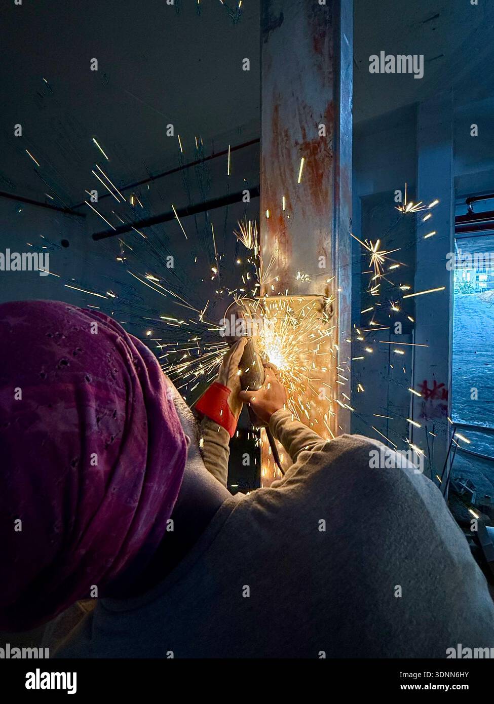 Worker in a pink turban using an angle grinder on a steel beam at a construction site, creating bright yellow sparks in a gritty industrial interior. - Smartphone Captured Stock Image