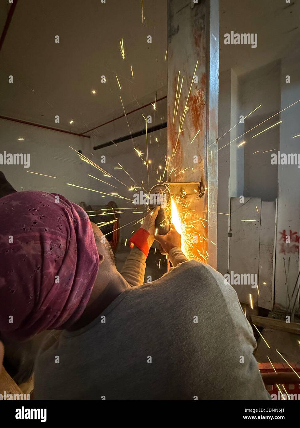 Worker in a pink turban using an angle grinder on a steel beam at a construction site, creating bright yellow sparks in a gritty industrial interior. - Smartphone Captured Stock Image