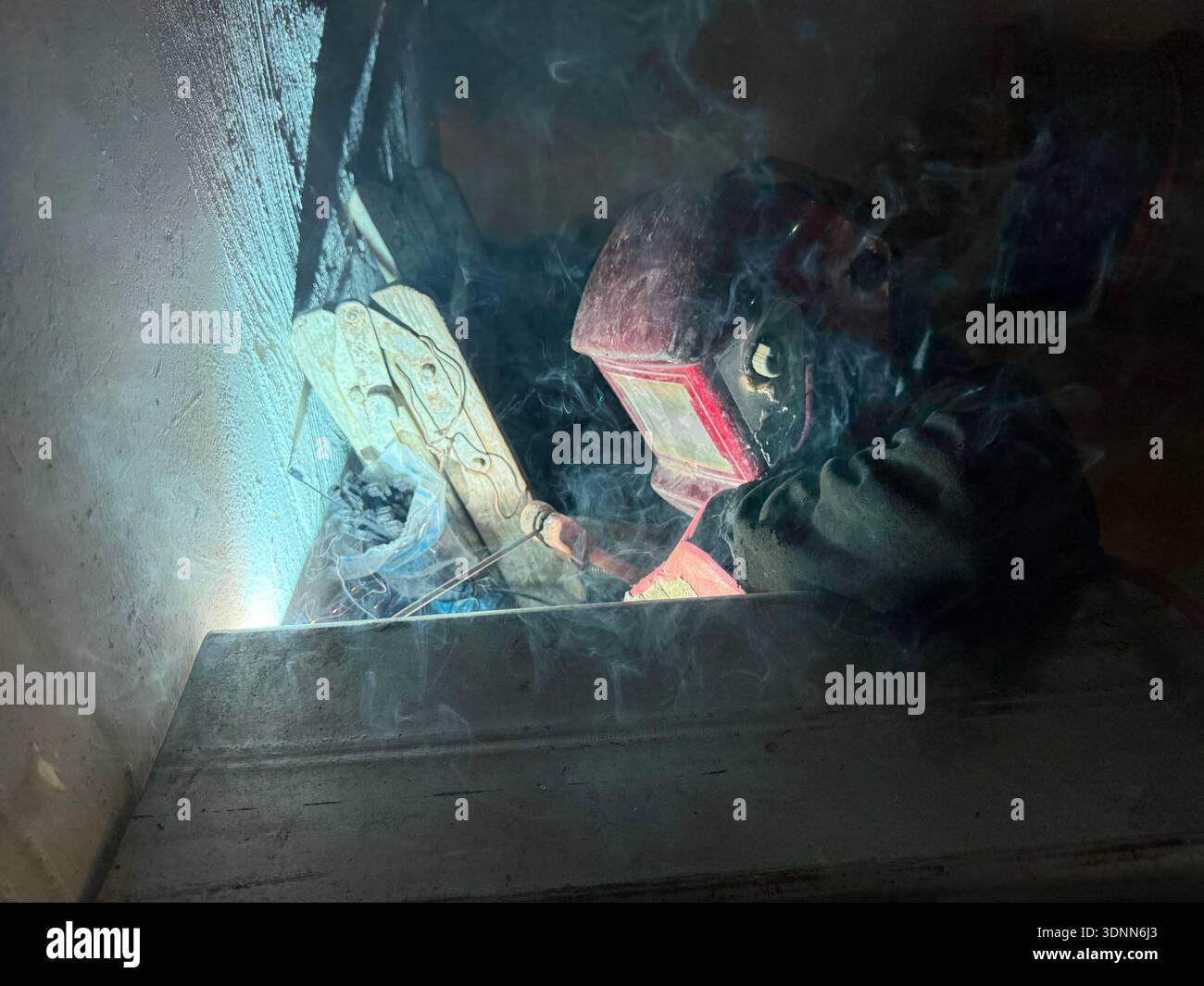 A professional welder in a protective red mask and gloves works in a dark, cramped space, creating bright arcs of light and smoke during fabrication. - Smartphone Captured Stock Image