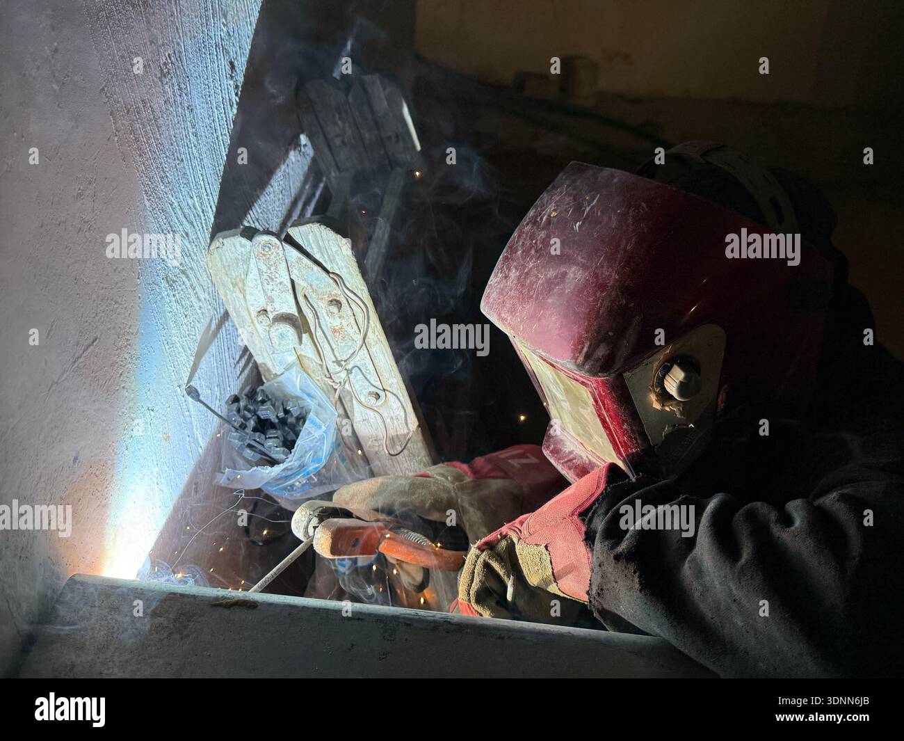 A professional welder in a protective red mask and gloves works in a dark, cramped space, creating bright arcs of light and smoke during fabrication. - Smartphone Captured Stock Image