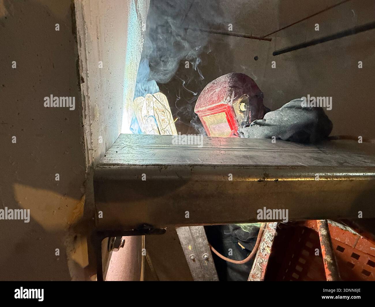 A professional welder in a protective red mask and gloves works in a dark, cramped space, creating bright arcs of light and smoke during fabrication. - Smartphone Captured Stock Image