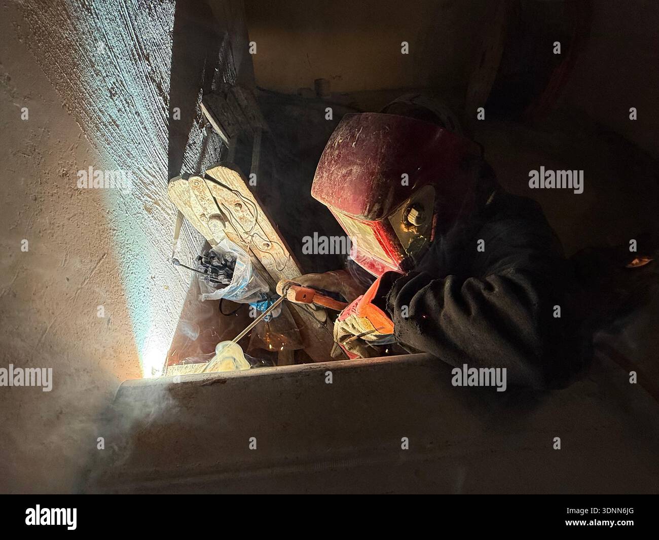 A professional welder in a protective red mask and gloves works in a dark, cramped space, creating bright arcs of light and smoke during fabrication. - Smartphone Captured Stock Image