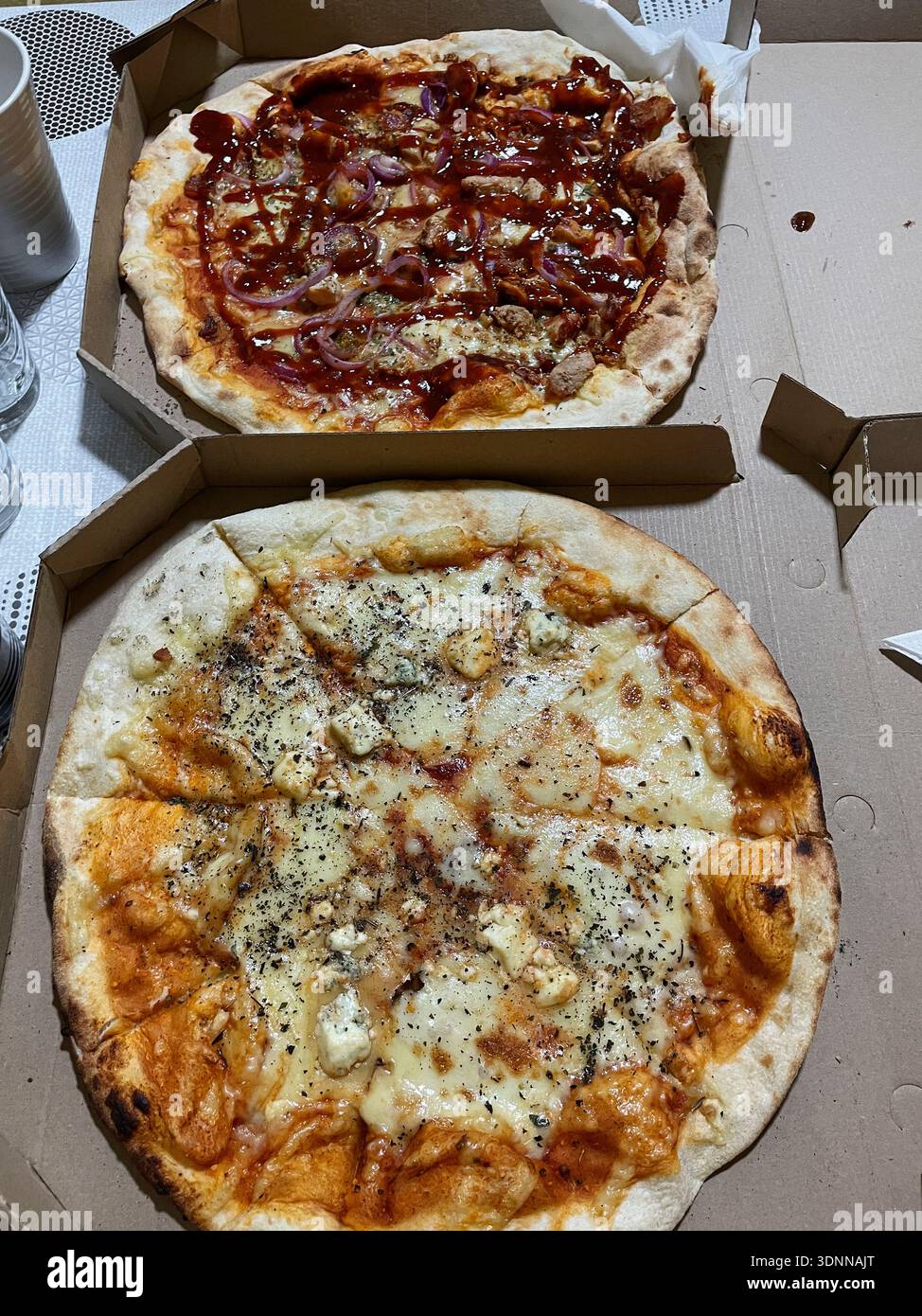 Two freshly baked pizzas served in open cardboard boxes on a table. One pizza topped with meat, onion, and sauce, the other with melted cheese and Stock Photo