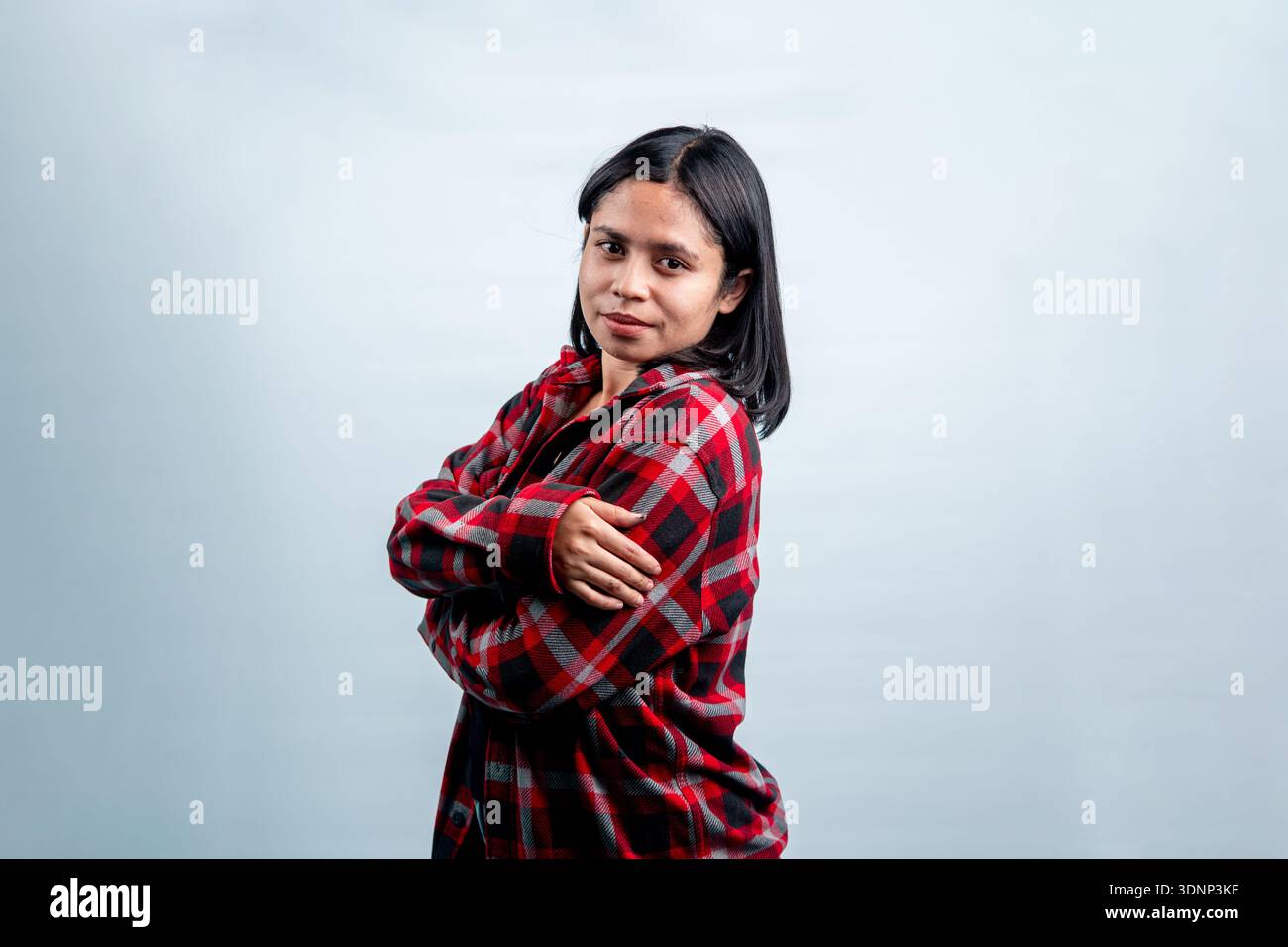 A charming young Southeast Asian woman smiling shyly at the camera, posing with folded arms in a casual red flannel shirt. Stock Photo