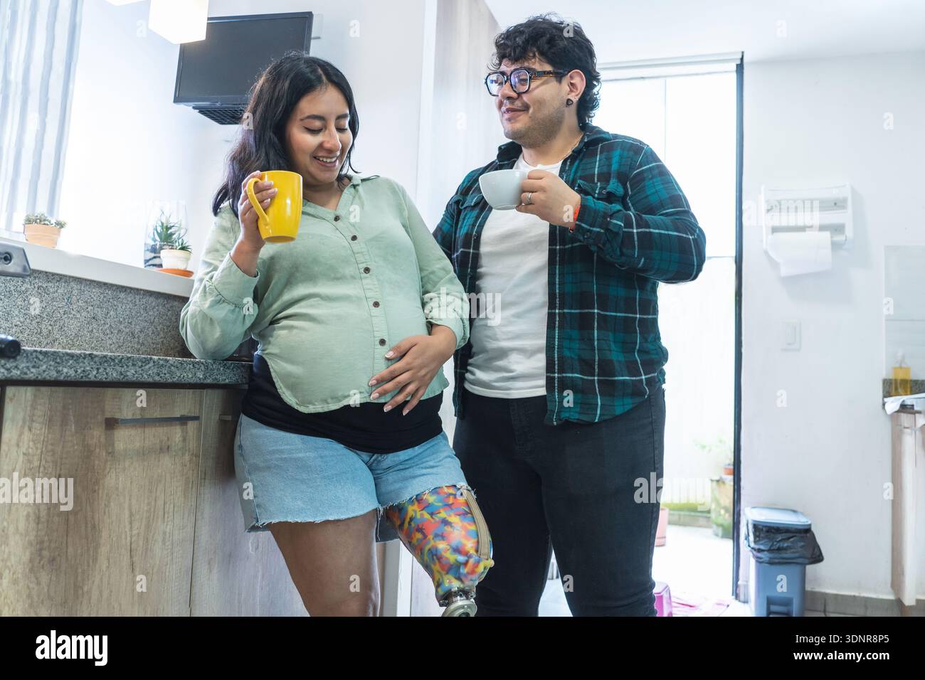 Pregnant woman with prosthetic leg embracing her baby bump beside her partner in the kitchen Stock Photo