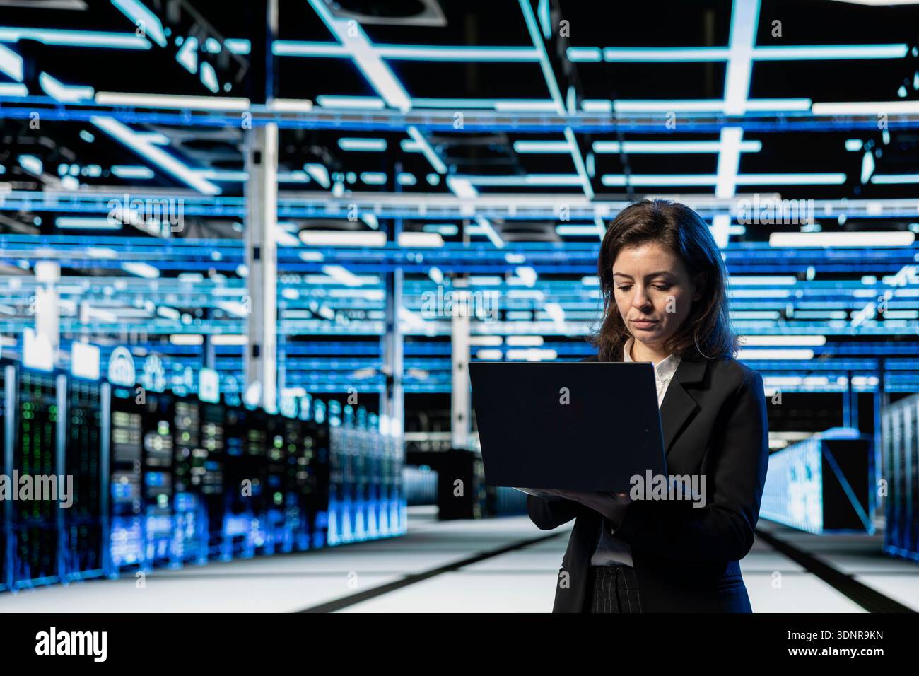 Data center engineer using laptop to do maintenance on cloud computing gear, VFX compositing overlay. Server farm worker walking between blue wireframe rackmounts using notebook Stock Photo