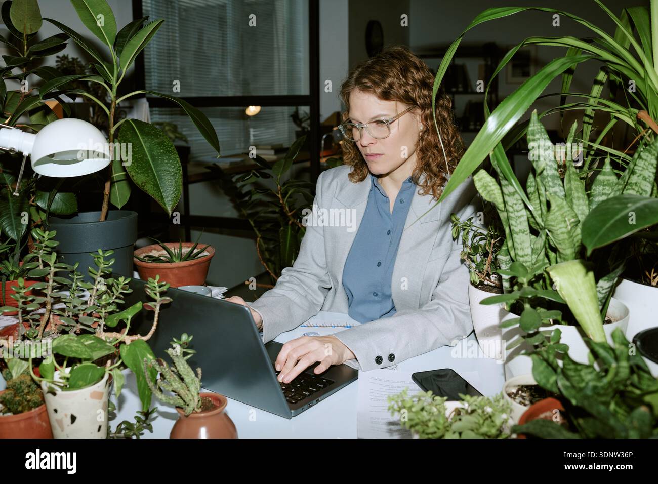 Young Woman Working Remotely on Laptop in Office Surrounded by Plants Stock Photo